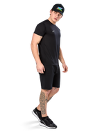 Mężczyzna z tatuażami w koszulce Alpinestars Point Performance Tee, czarny t-shirt techniczny z krótkim rękawem, małe białe logo, w zestawie z czarnymi szortami, czarną czapką z kolorową grafiką i czarno-białymi sneakersami, sportowy styl lifestyle