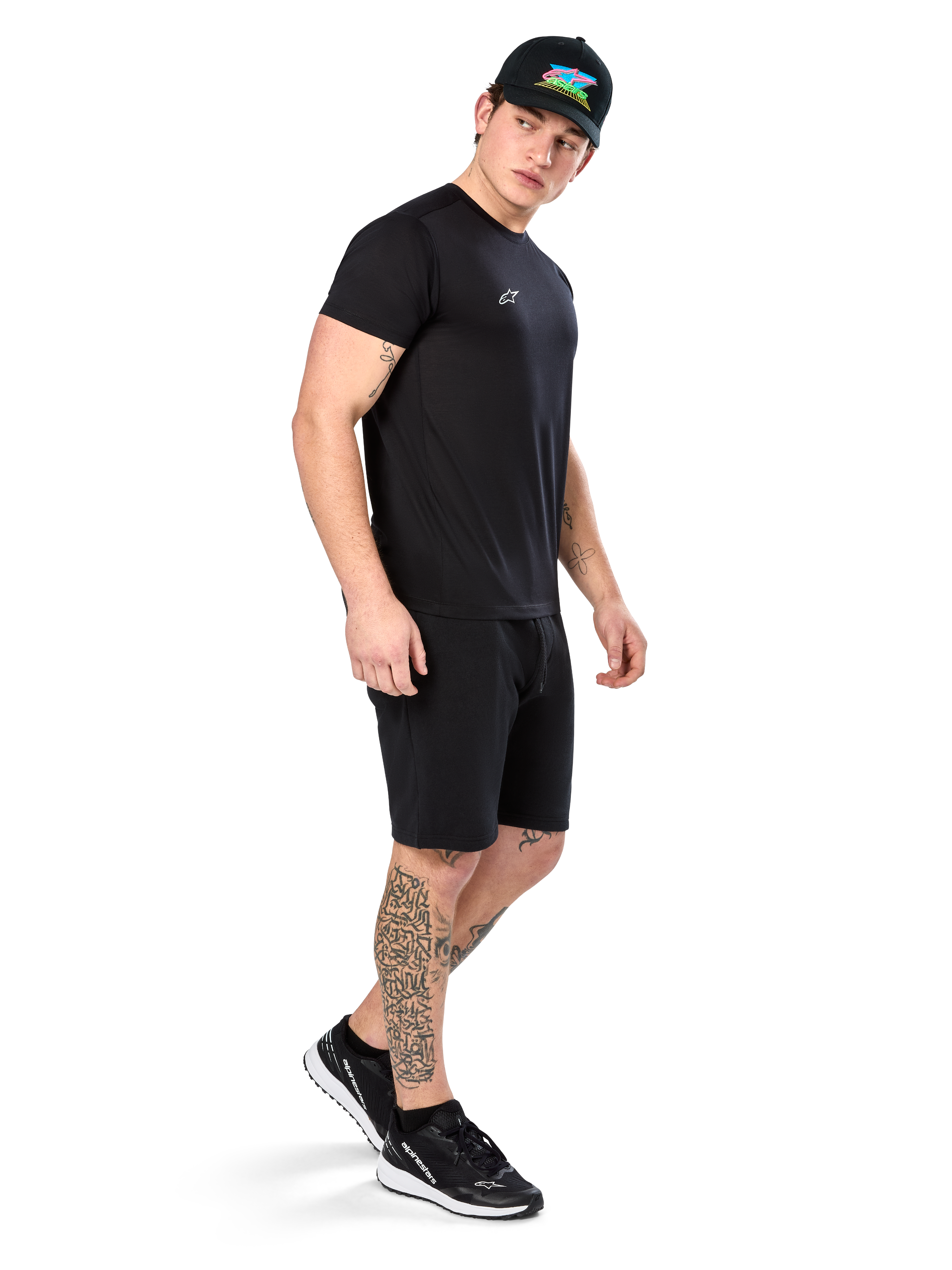 Mężczyzna z tatuażami w koszulce Alpinestars Point Performance Tee, czarny t-shirt techniczny z krótkim rękawem, małe białe logo, w zestawie z czarnymi szortami, czarną czapką z kolorową grafiką i czarno-białymi sneakersami, sportowy styl lifestyle