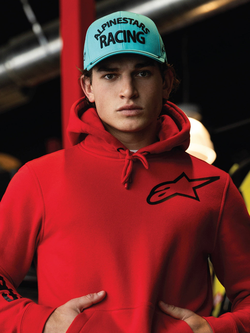 Czapka Alpinestars Luminary, jasnoniebieska czapka typu snapback z zakrzywionym daszkiem, czarny haft 3D z napisem „ALPINESTARS RACING”, noszona z jaskrawoczerwoną bluzą z kapturem Alpinestars, codzienna odzież uliczna w stylu motorsport