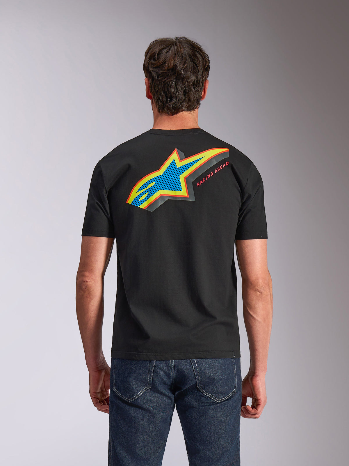 Alpinestars Angler CSF Tee, koszulka z krótkim rękawem, czarna, widok z tyłu przedstawiający wyraziste wielokolorowe logo Astars z perforowanym wypełnieniem i napisem „RACING AHEAD”, niebieskie jeansy, codzienna odzież inspirowana sportami motorowymi