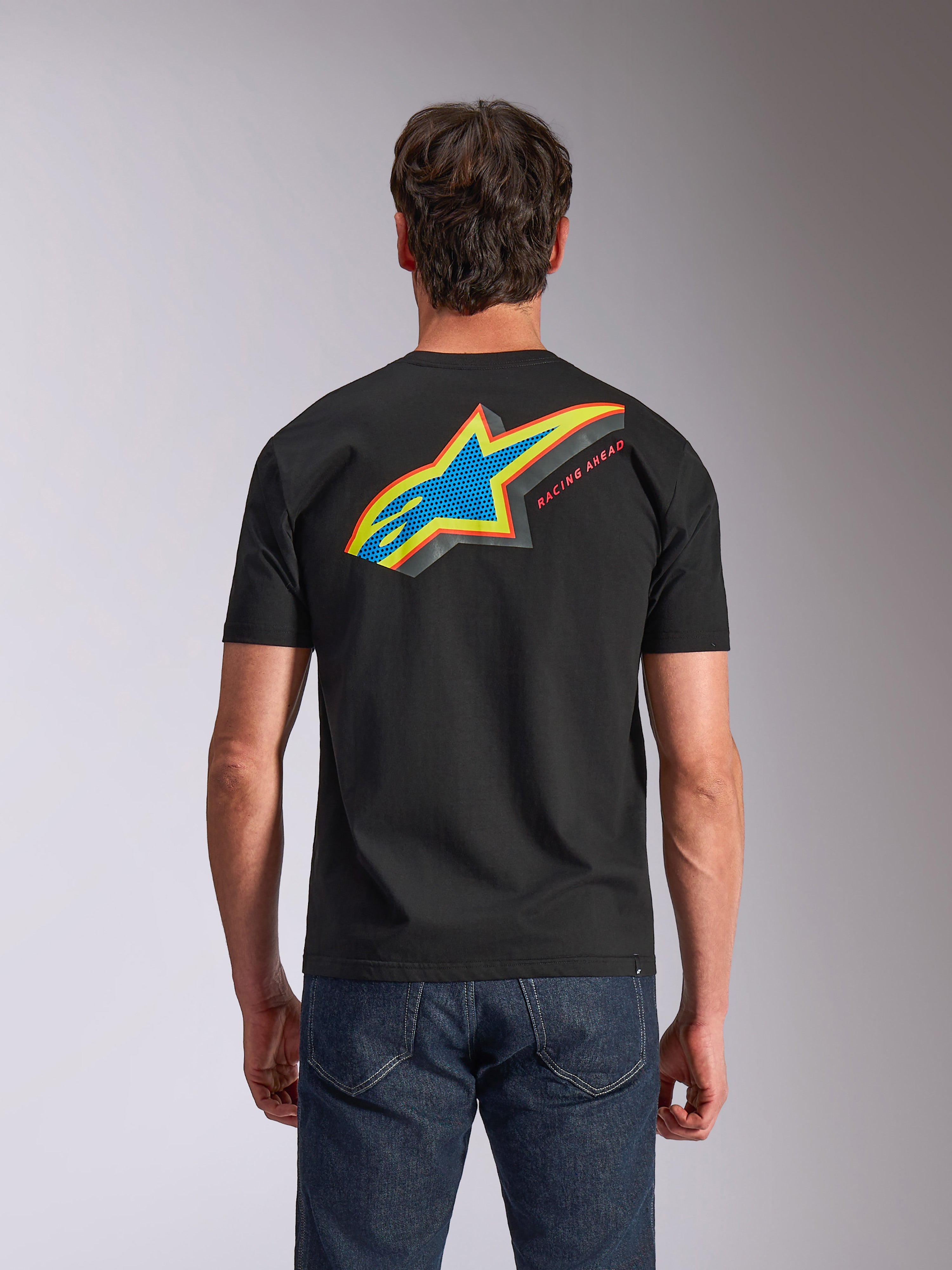Alpinestars Angler CSF Tee, koszulka z krótkim rękawem, czarna, widok z tyłu przedstawiający wyraziste wielokolorowe logo Astars z perforowanym wypełnieniem i napisem „RACING AHEAD”, niebieskie jeansy, codzienna odzież inspirowana sportami motorowymi