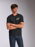 Alpinestars Angler CSF Tee, t-shirt z krótkim rękawem, czarny, z wyrazistym, wielokolorowym logo Astars na piersi, noszony przez modela ze skrzyżowanymi ramionami w niebieskich jeansach, codzienna odzież uliczna inspirowana motorsportem