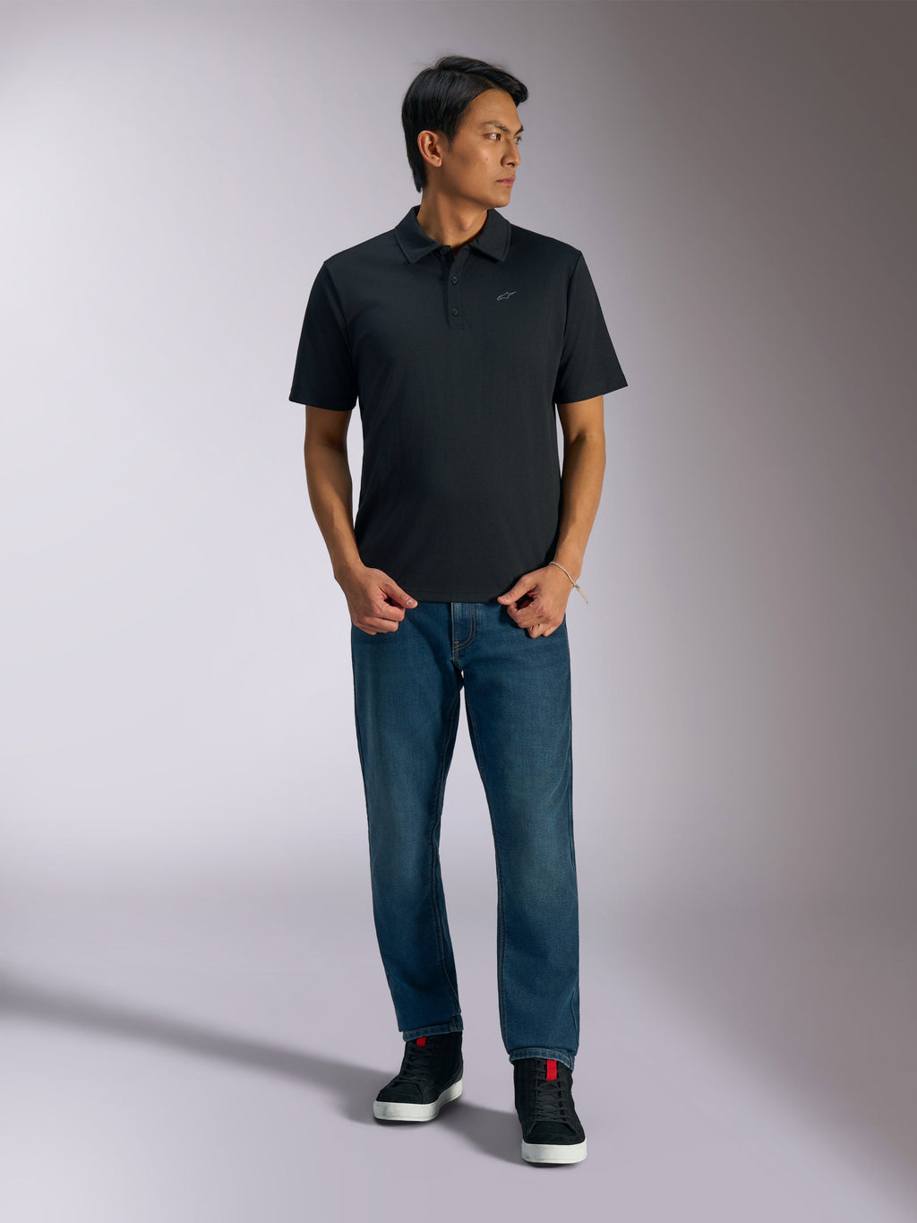 Alpinestars Impetus Sport Polo, czarna koszulka polo z krótkim rękawem, zapięcie na trzy guziki i szare logo Astars na piersi, model w niebieskich jeansach i czarnych wysokich sneakersach, casualowa odzież sportowa inspirowana sportami motorowymi