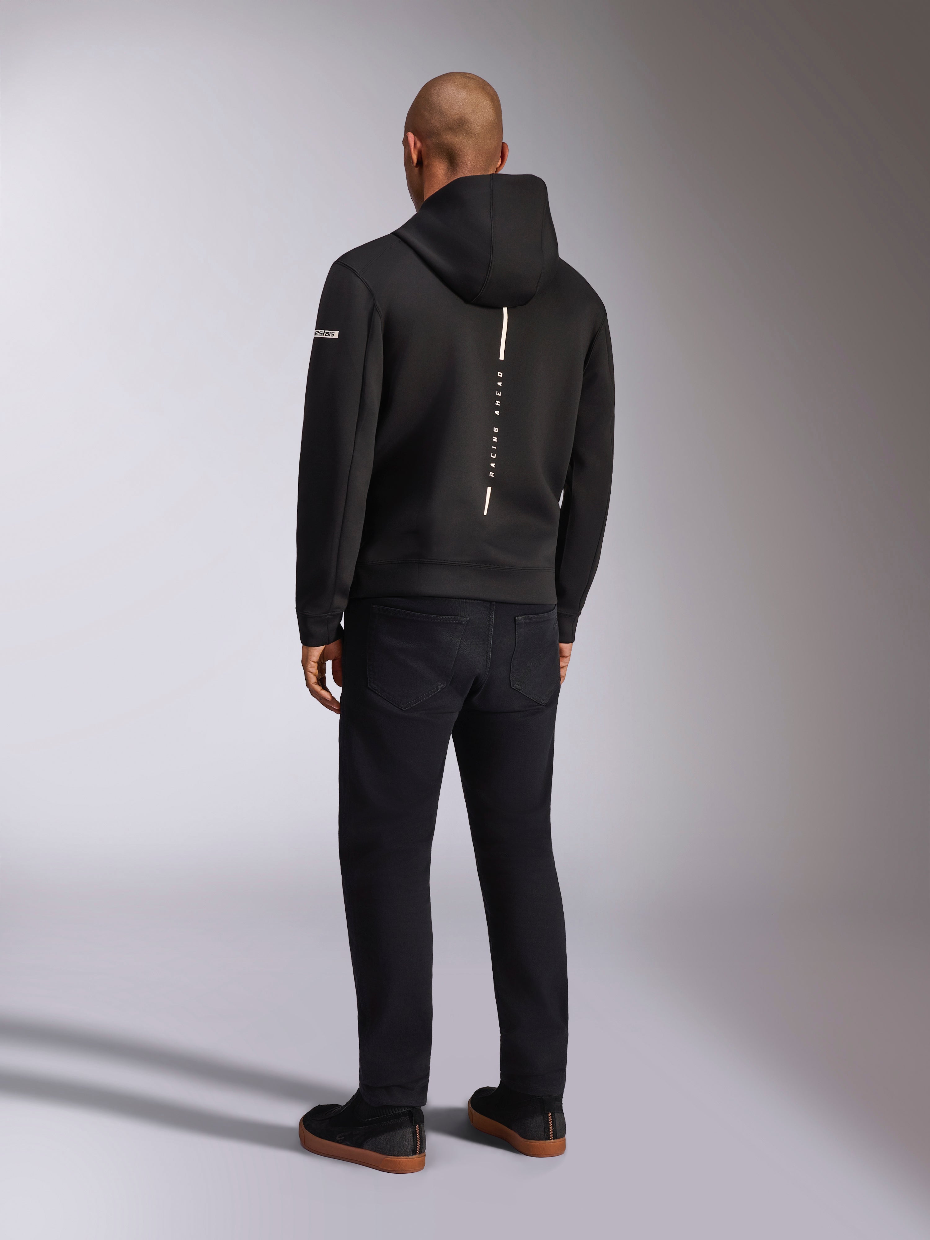 Alpinestars Insight Hoodie, czarna sportowa bluza z kapturem, widok z tyłu przedstawiający pionowy biały pasek z brandingiem „RACING AHEAD”, logo na rękawie, w zestawieniu z czarnymi spodniami i sneakersami, casualowy design inspirowany sportami motorowymi
