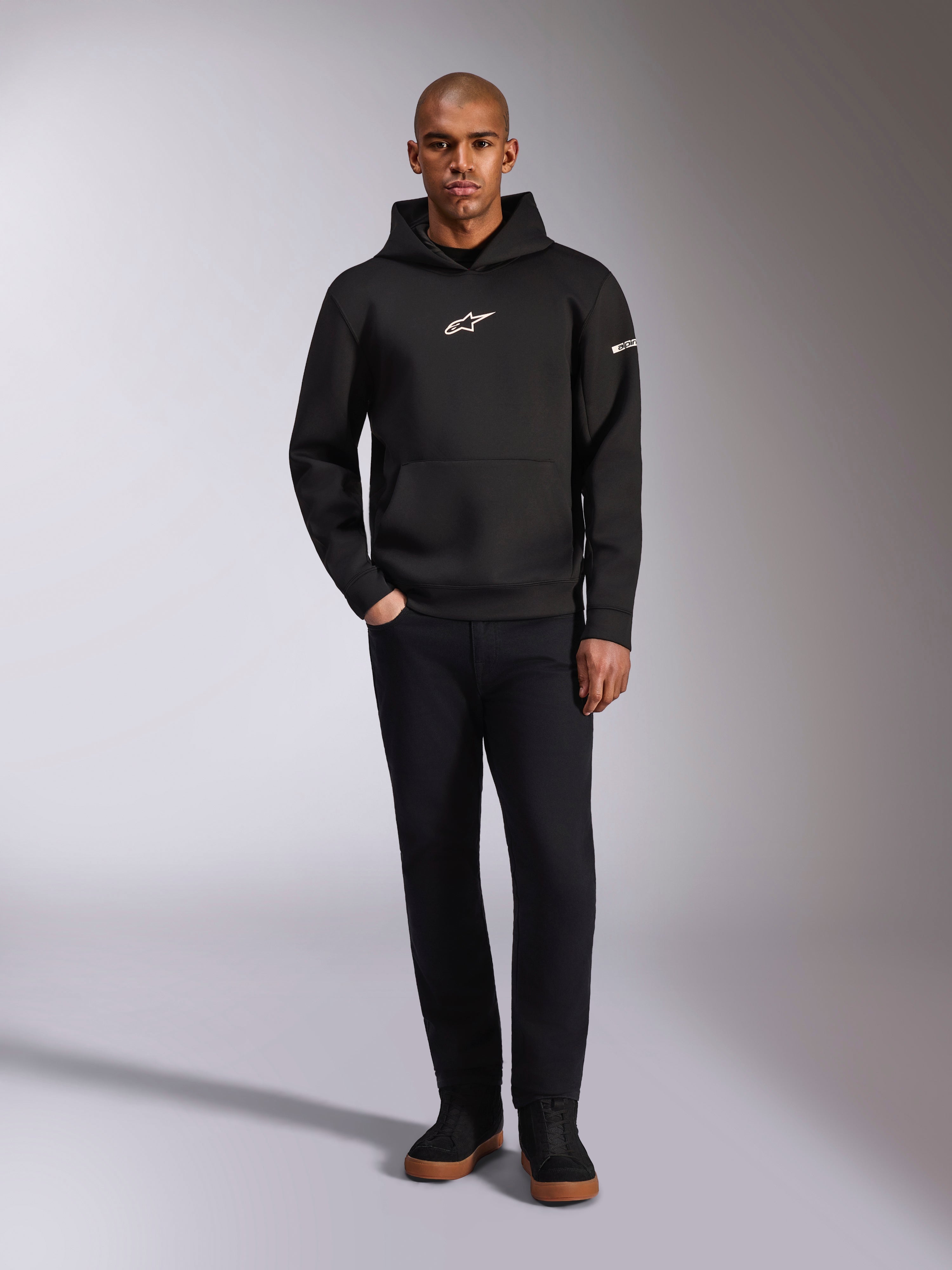 Alpinestars Insight Hoodie, czarna bluza z kapturem typu pullover z białym logo Astars na piersi i rękawie, prezentowana na modelu w czarnych spodniach i czarnych sneakersach, casualowy streetwear inspirowany motorsportem