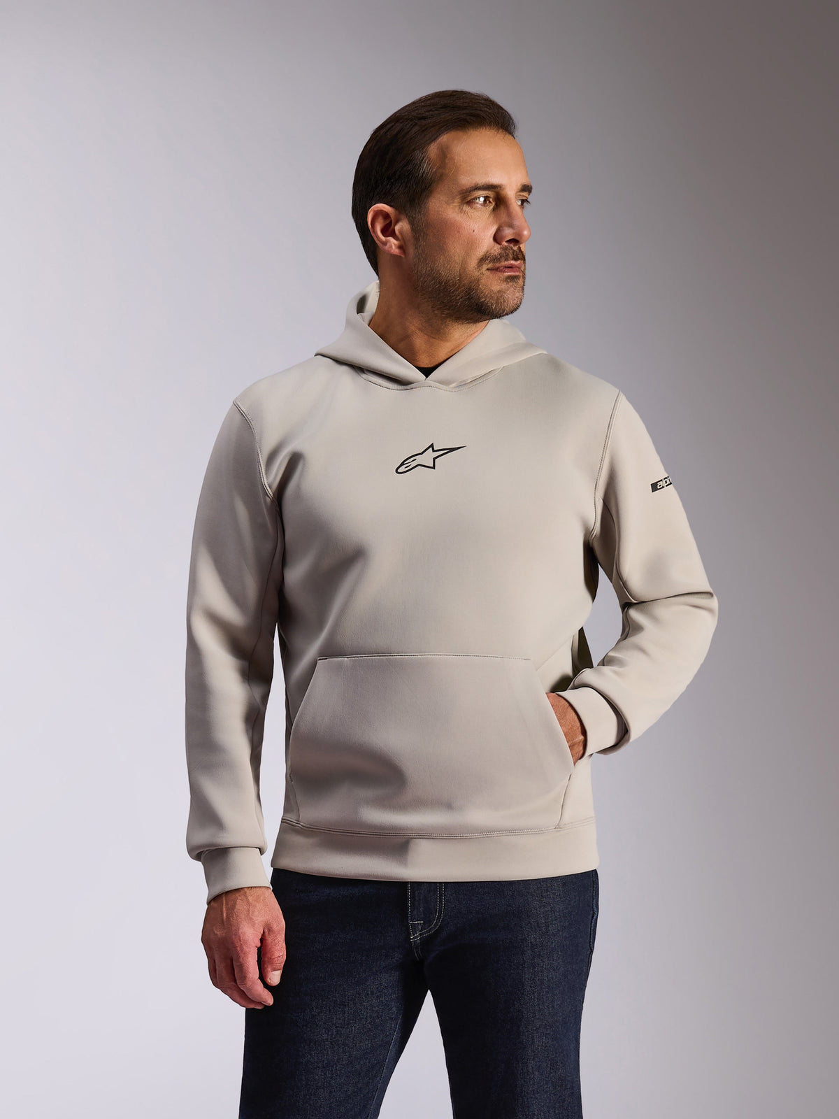 Alpinestars Insight Hoodie, sportowa odzież codzienna, kolor szary Cement, fason typu pullover z kieszenią kangurką i małym czarnym logo Astars na piersi, długie rękawy z napisem Alpinestars na lewym ramieniu, prezentowana przez modela w ciemnych jeansach, lifestylowa odzież motorsportowa