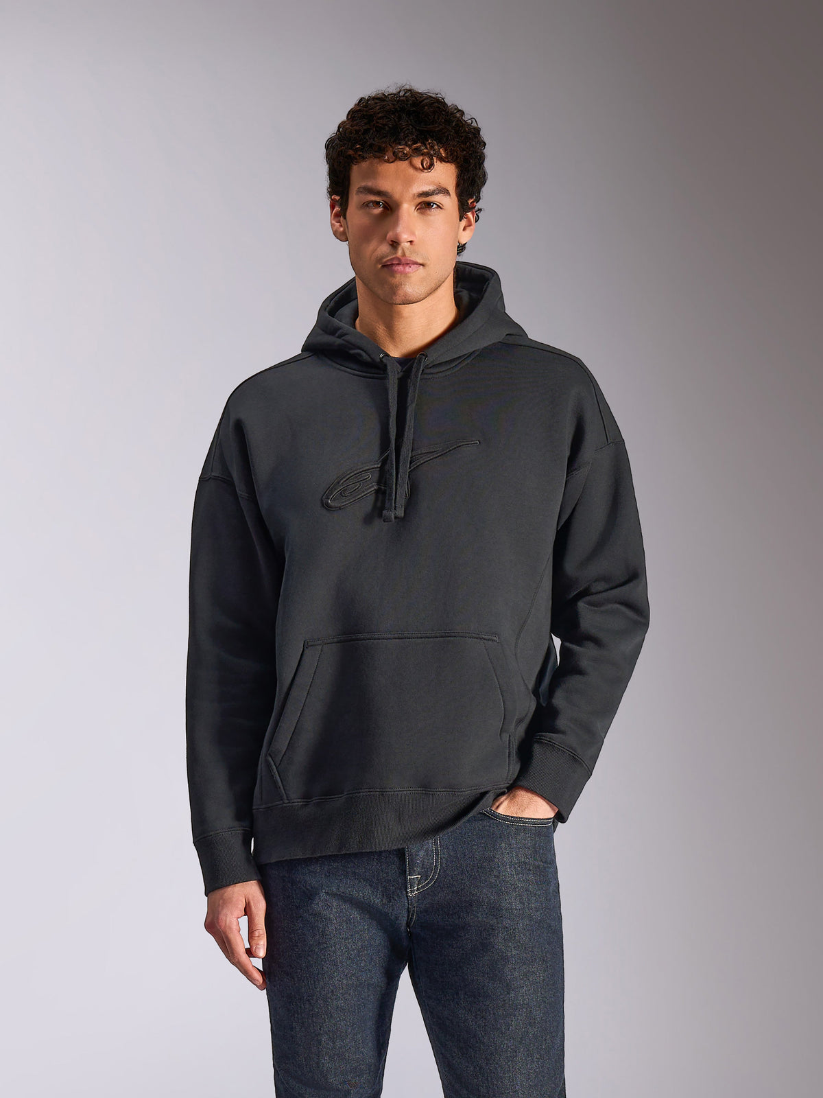 Alpinestars Emergent Oversized Hoodie, Black, male model wearing oversized pullover fleece hoodie, tonal 3D embroidered Astars logo, kangaroo pocket, drawstring hood, casual motorsport-inspired streetwear