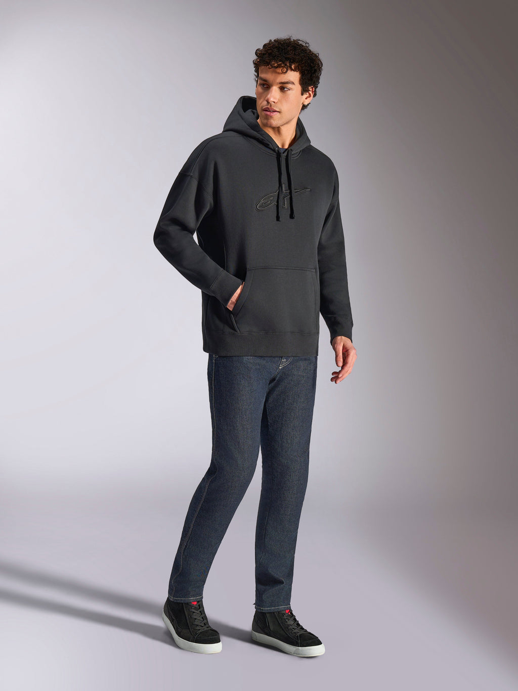Alpinestars Emergent Oversized Hoodie, czarna bluza streetwearowa, tonalne haftowane logo 3D Astars na piersi, kieszeń kangurka, oversizowy krój, model w ciemnoniebieskich dżinsach i czarnych sneakersach