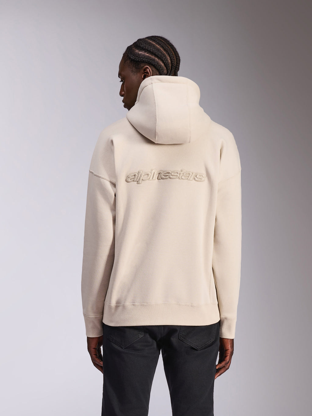 Alpinestars Emergent Oversized Hoodie, casualowa bluza streetwearowa, kolor piaskowy złamana biel, haftowane logo 3D Alpinestars na plecach, krój oversize z prążkowanym dołem i mankietami, widok z tyłu na modelu w czarnych spodniach.