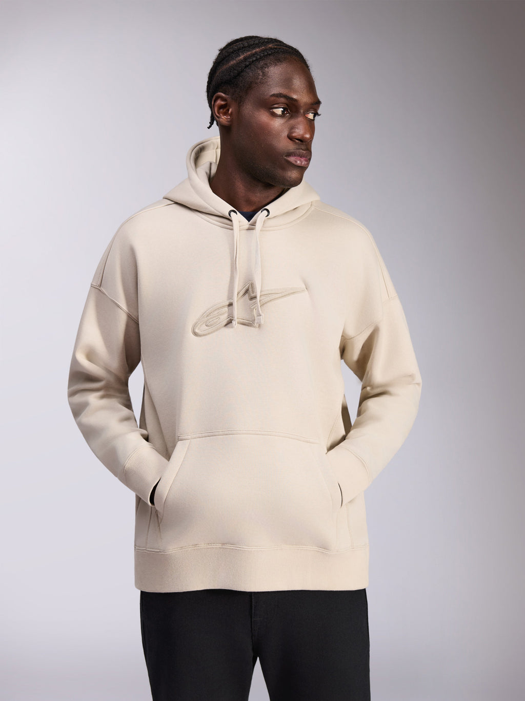 Alpinestars Emergent Oversized Hoodie, kolor piaskowy, bluza o luźnym kroju z haftowanym logo 3D Astars w tym samym odcieniu na piersi, kieszeń kangurka, kaptur ze sznurkami, styl streetwear prezentowany przez modela w czarnych spodniach.