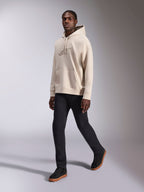Alpinestars Emergent Oversized Hoodie, casualowy streetwear, piaskowy odcień bieli, tonalne haftowane logo 3D, model w czarnych spodniach i czarnych sneakersach, luźny krój oversize z kieszenią kangurka i kapturem ze sznurkiem