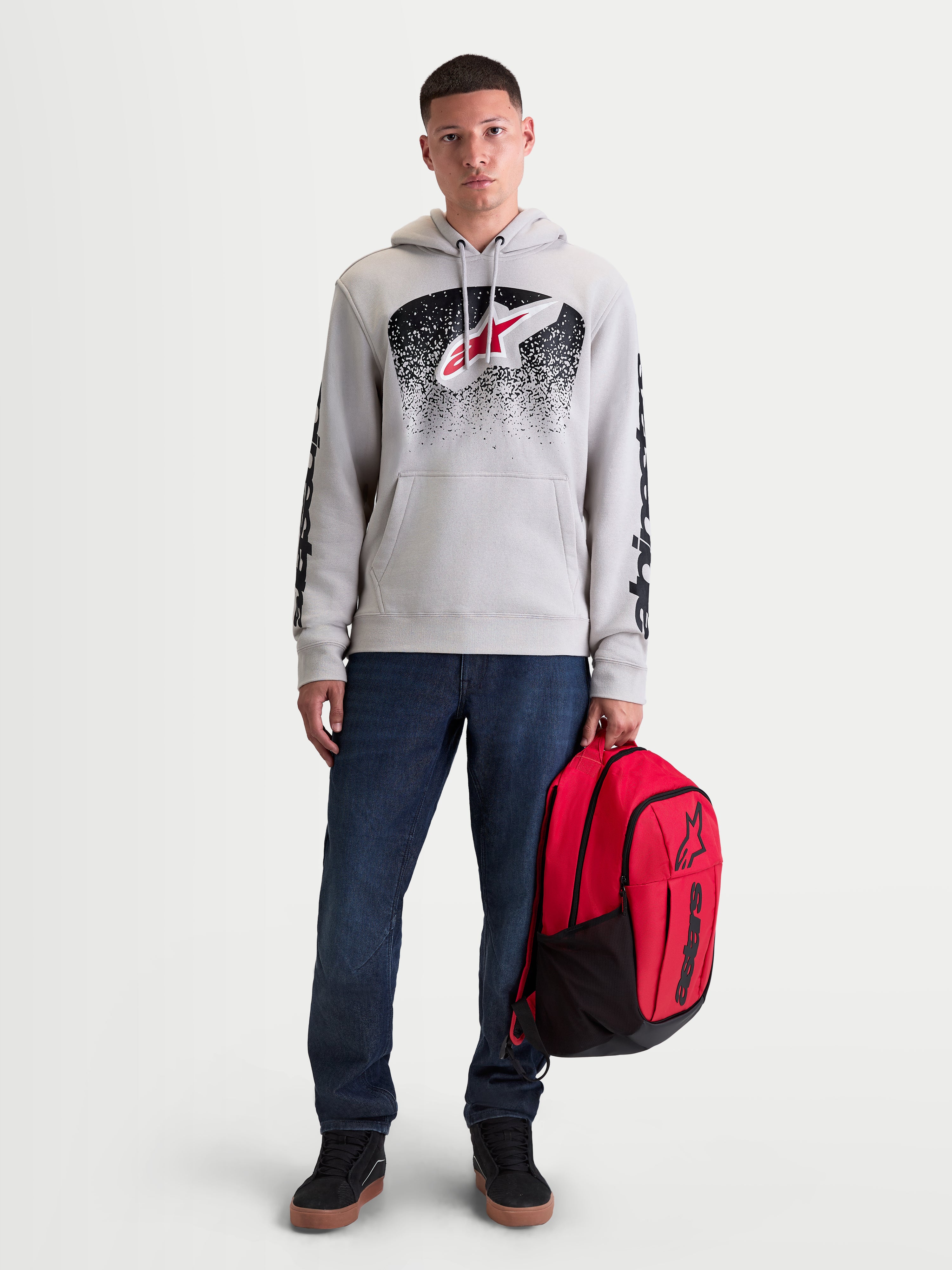 Alpinestars Bluza z kapturem Obsessed, męska bluza polarowa sportswear, szara z czerwono-czarną grafiką inspirowaną MX, logo na rękawach, model trzymający czerwony plecak Alpinestars.