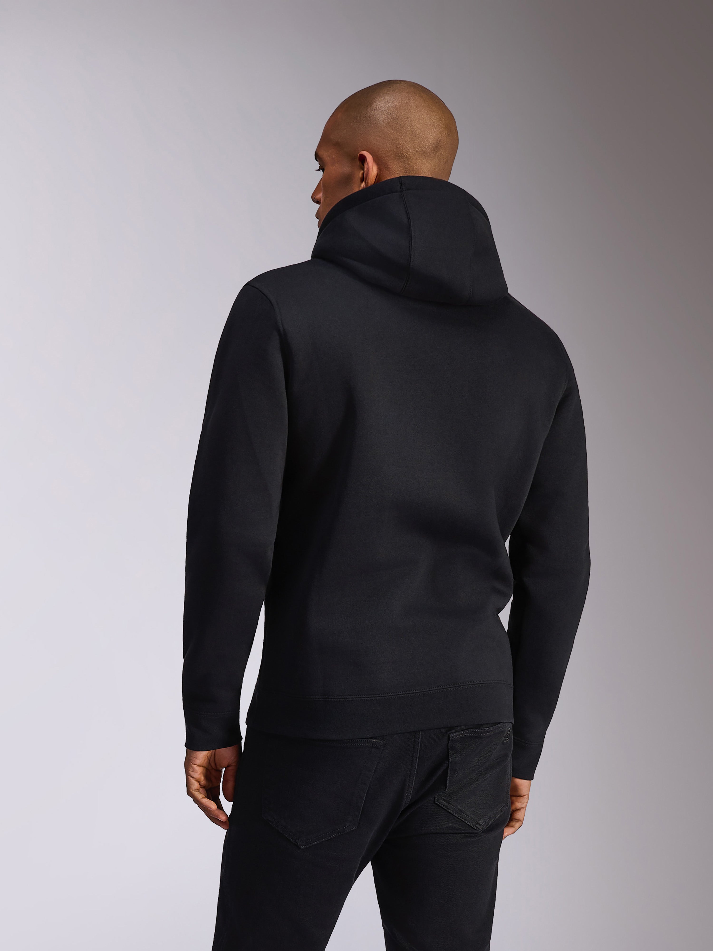 Alpinestars Ellipsoid Hoodie, czarna, widok z tyłu osoby ubranej w wkładaną przez głowę bluzę z miękkiego polaru z kapturem, długie rękawy z żebrowanymi mankietami i dołem, stylizacja z czarnymi dżinsami, codzienna odzież sportowa inspirowana sportami motorowymi