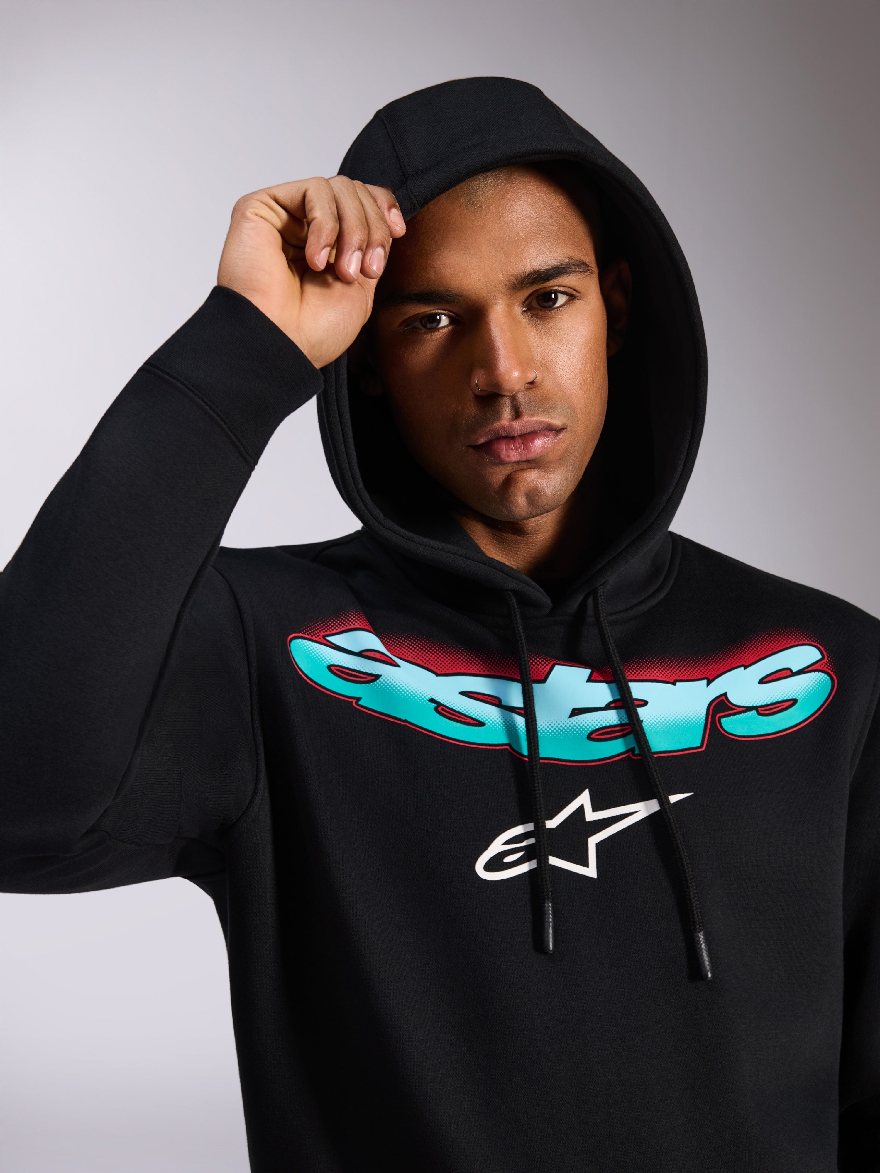 Alpinestars Ellipsoid Hoodie, czarna bluza z polaru z kapturem, stylizowany turkusowo-czerwony napis Astars i białe logo na piersi, noszona przez modela z założonym kapturem, codzienna odzież uliczna inspirowana sportami motorowymi