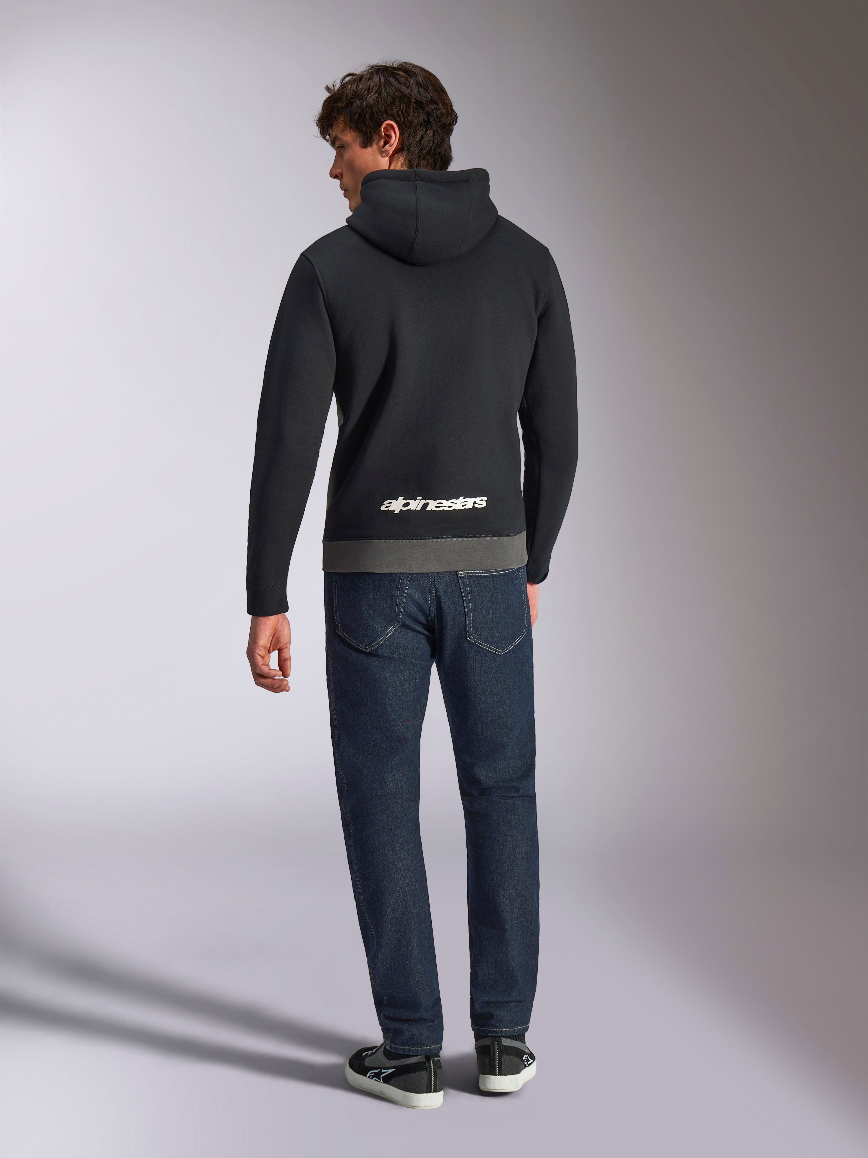 Alpinestars Parallel Hoodie, rear view of a man wearing a black and gunmetal color-blocked pullover fleece, white Alpinestars word logo on the lower back, paired with dark blue jeans, casual motorsport-inspired streetwear