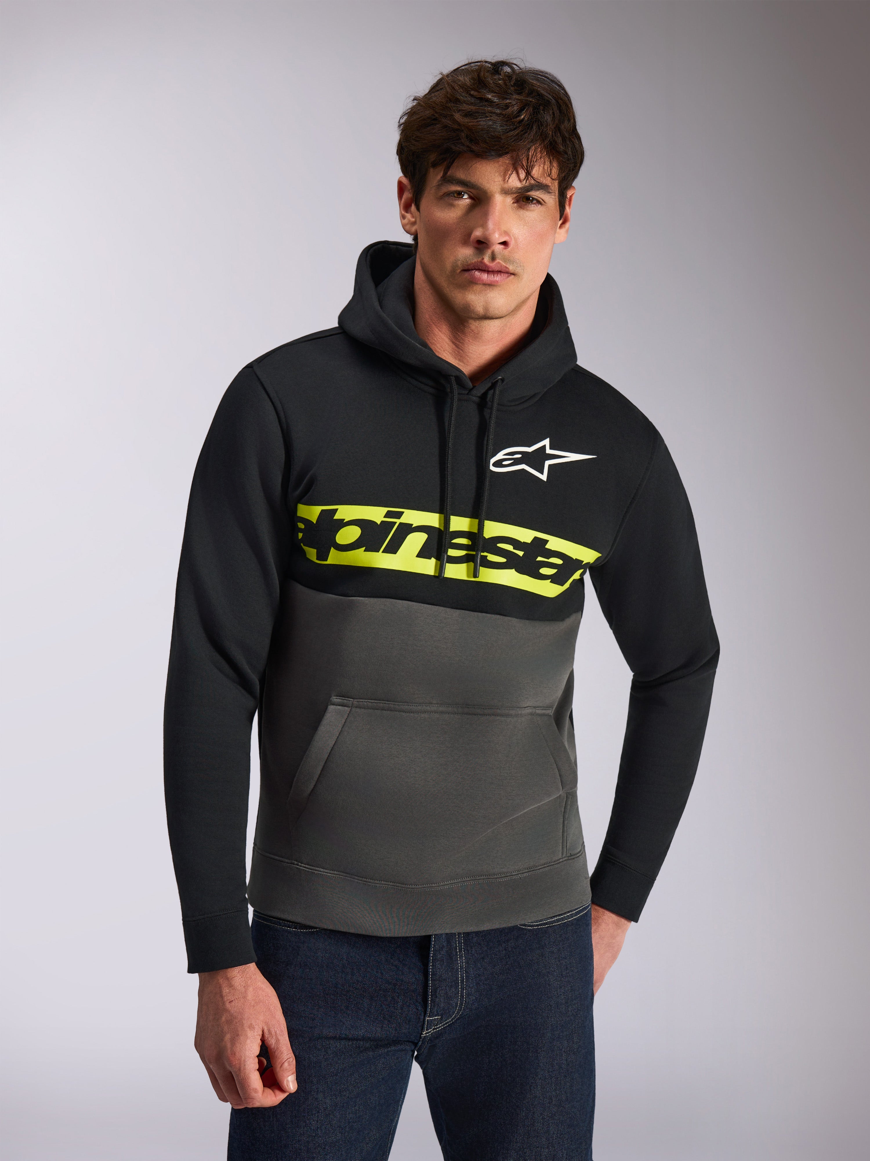 Alpinestars Parallel Hoodie, casual streetwear, color-blocked black and gunmetal grey, bright yellow chest stripe with Alpinestars word logo, white Astars logo, worn by a model with blue jeans, modern motorsport-inspired design