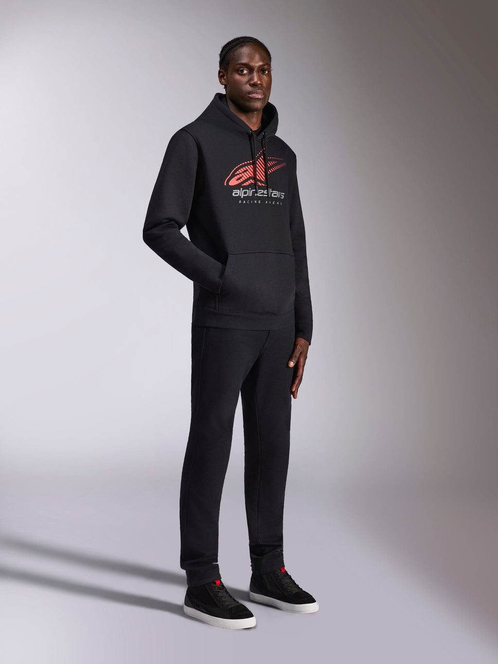 Alpinestars Pulse Hoodie, czarna bluza z kapturem typu pullover, z czerwono-białym logo Astars z efektem cieniowania i napisem „Racing Ahead”, w zestawie z pasującymi czarnymi spodniami i sneakersami, codzienna odzież streetwear inspirowana motorsportem.