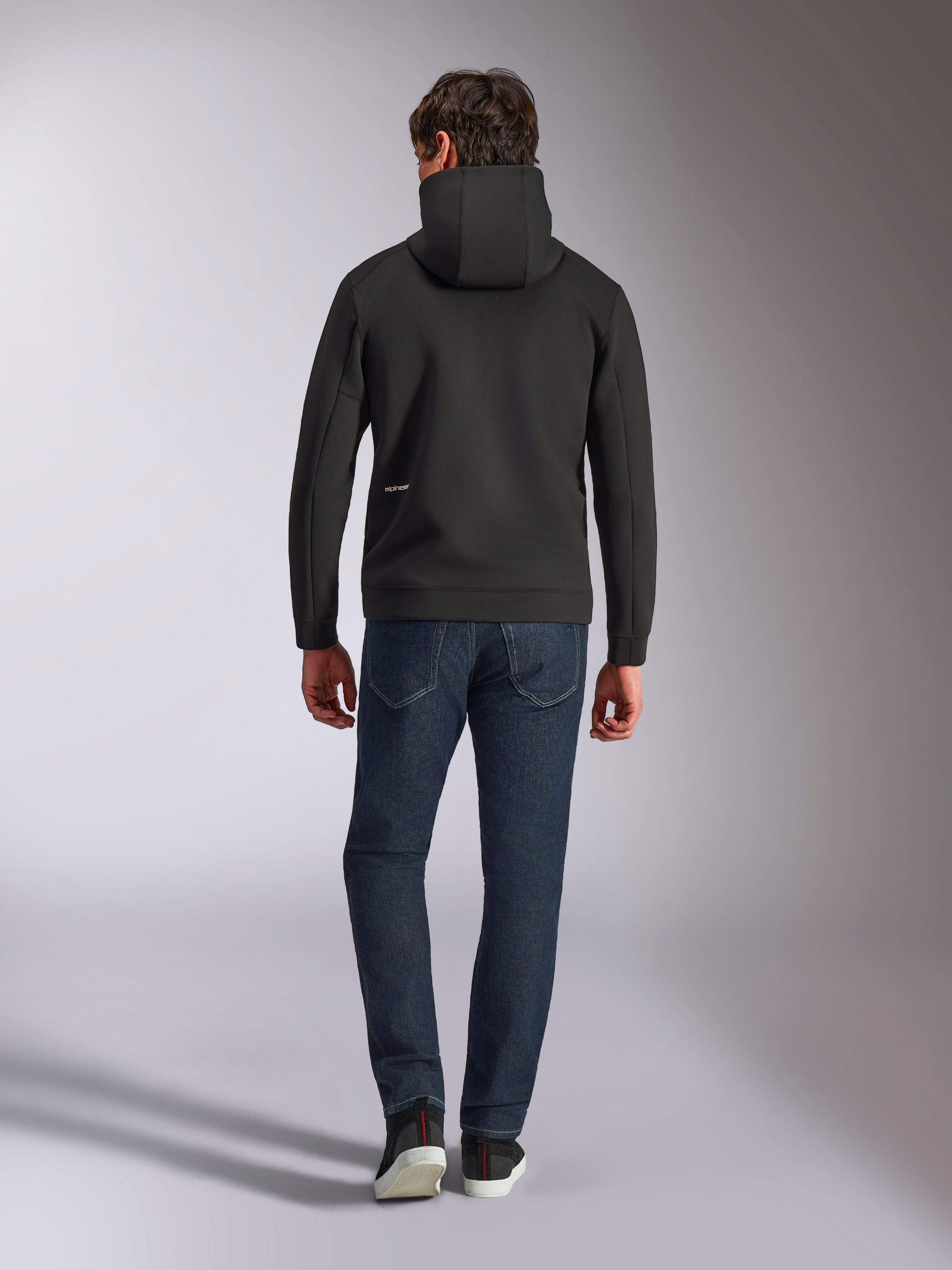 Alpinestars Cypher Hoodie, czarny dwustronny polar, widok z tyłu z małym białym logo Alpinestars na lewym biodrze, wysoki kaptur, połączona z ciemnoniebieskimi jeansami, casualowy streetwear inspirowany motorsportem