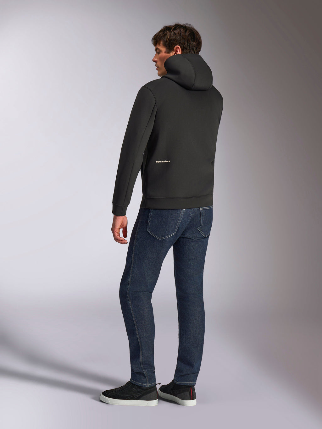 Alpinestars Cypher Hoodie, czarna codzienna bluza z kapturem zapinana na zamek, widok z tyłu ukazujący wysoki kaptur i białe logo Alpinestars w dolnej części pleców, zestawiona z ciemnoniebieskimi dżinsami i czarnymi tenisówkami, nowoczesny design inspirowany sportami motorowymi