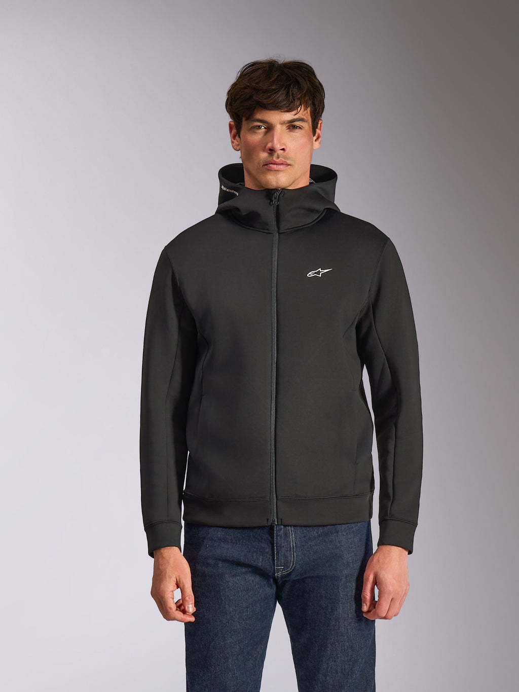 Alpinestars Cypher Hoodie, czarna rozpinana bluza z kapturem z polaru, wysoki kołnierz z białym brandingiem i małe białe logo Astars na piersi, prezentowana przez modela w niebieskich jeansach, codzienna odzież streetwear inspirowana motorsportem.