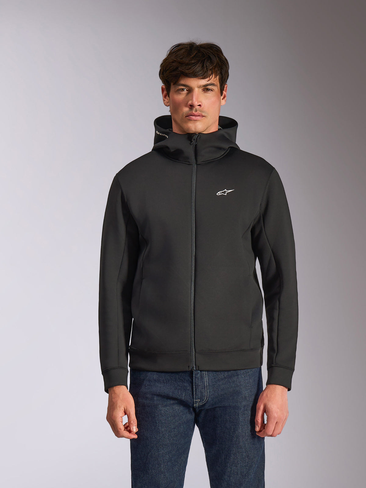 Alpinestars Cypher Hoodie, czarna rozpinana bluza z kapturem z polaru, wysoki kołnierz z białym brandingiem i małe białe logo Astars na piersi, prezentowana przez modela w niebieskich jeansach, codzienna odzież streetwear inspirowana motorsportem.