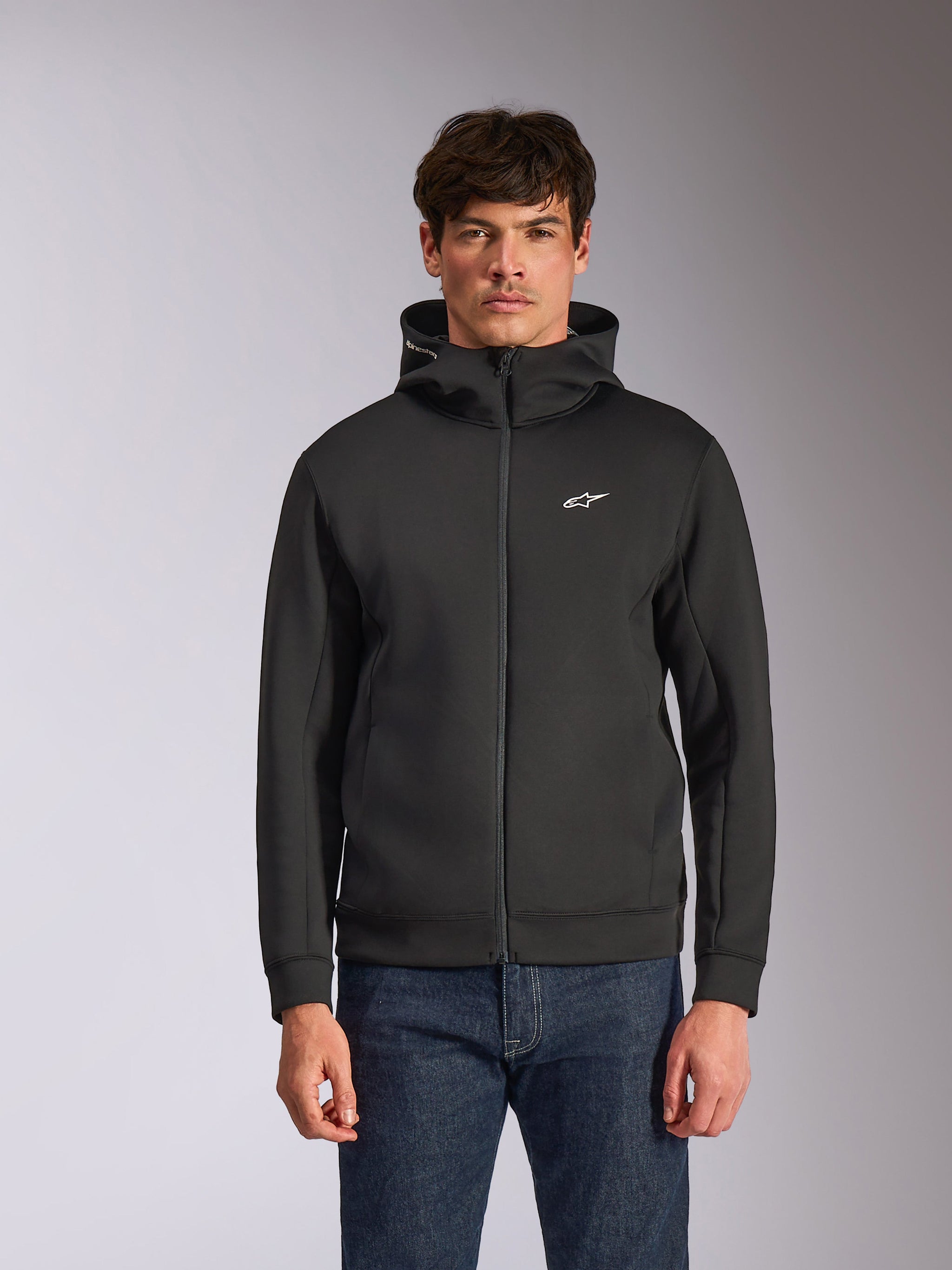 Alpinestars Cypher Hoodie, czarna rozpinana bluza z kapturem z polaru, wysoki kołnierz z białym brandingiem i małe białe logo Astars na piersi, prezentowana przez modela w niebieskich jeansach, codzienna odzież streetwear inspirowana motorsportem.