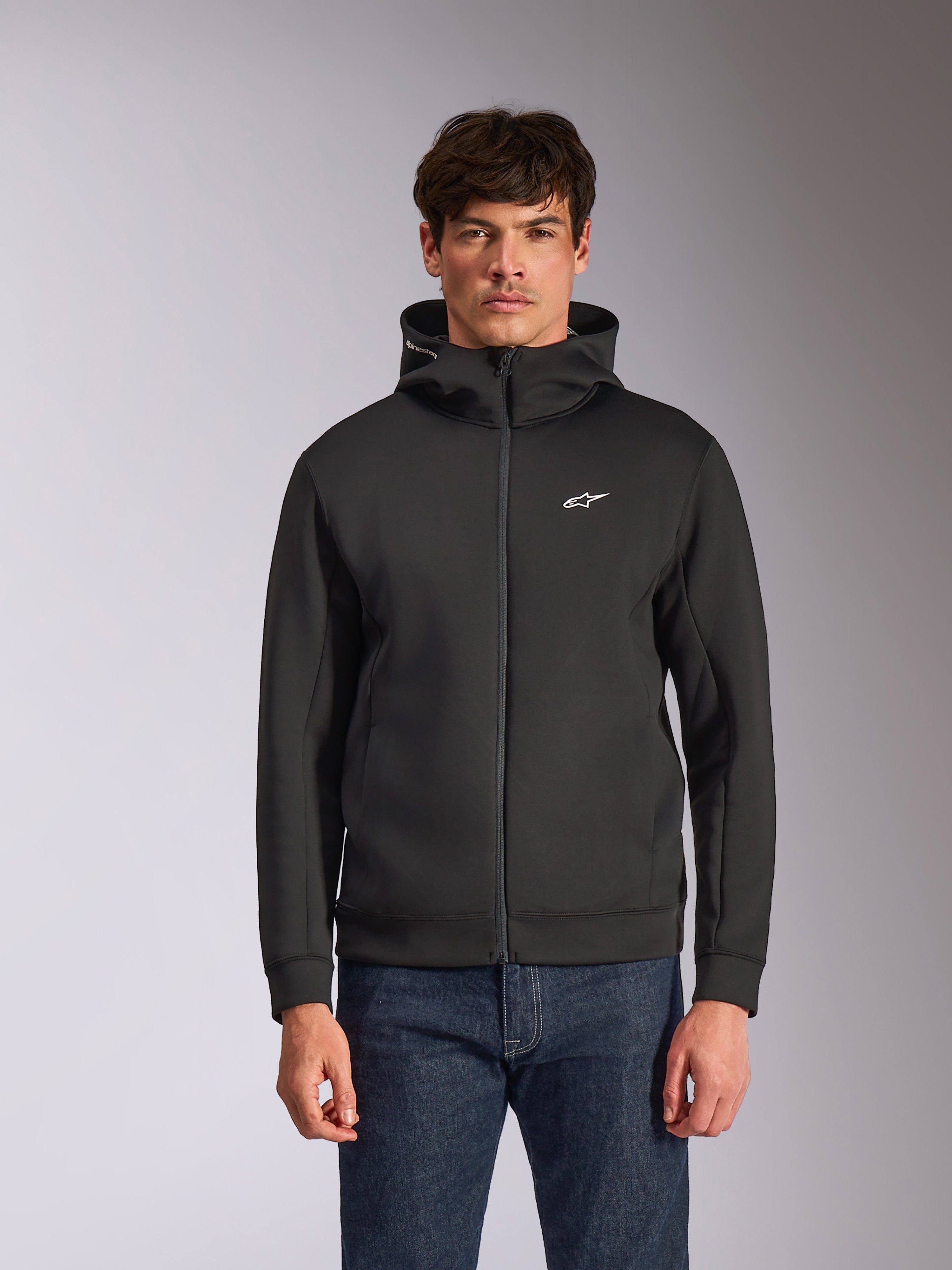 Alpinestars Cypher Hoodie, czarna rozpinana bluza z kapturem z polaru, wysoki kołnierz z białym brandingiem i małe białe logo Astars na piersi, prezentowana przez modela w niebieskich jeansach, codzienna odzież streetwear inspirowana motorsportem.