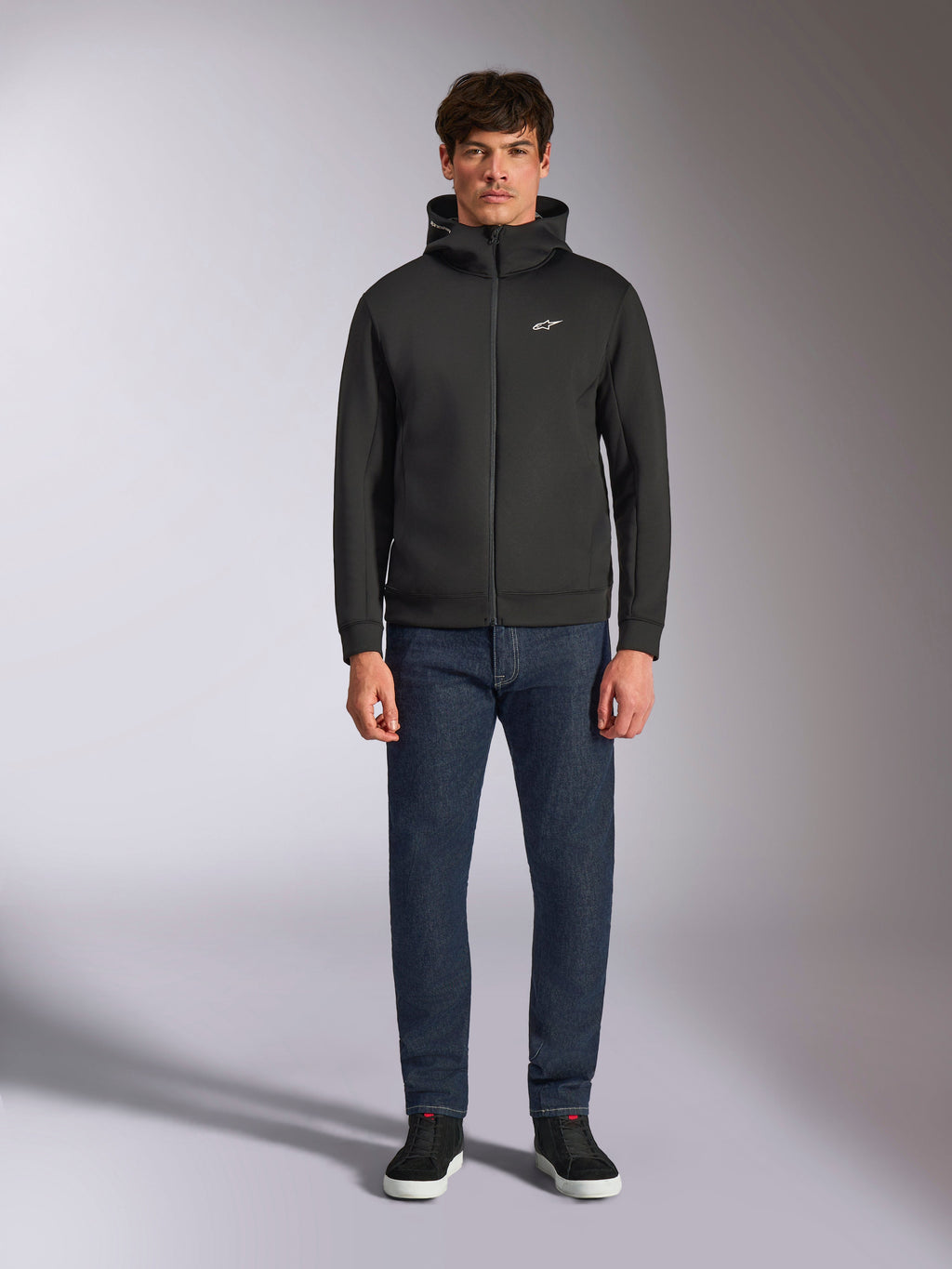 Model w bluzie Alpinestars Cypher Hoodie, czarna rozpinana bluza z polaru, wysoki kaptur z małym białym logo Astars na piersi, ciemne jeansy i czarne sneakersy, casualowy streetwear inspirowany motorsportem