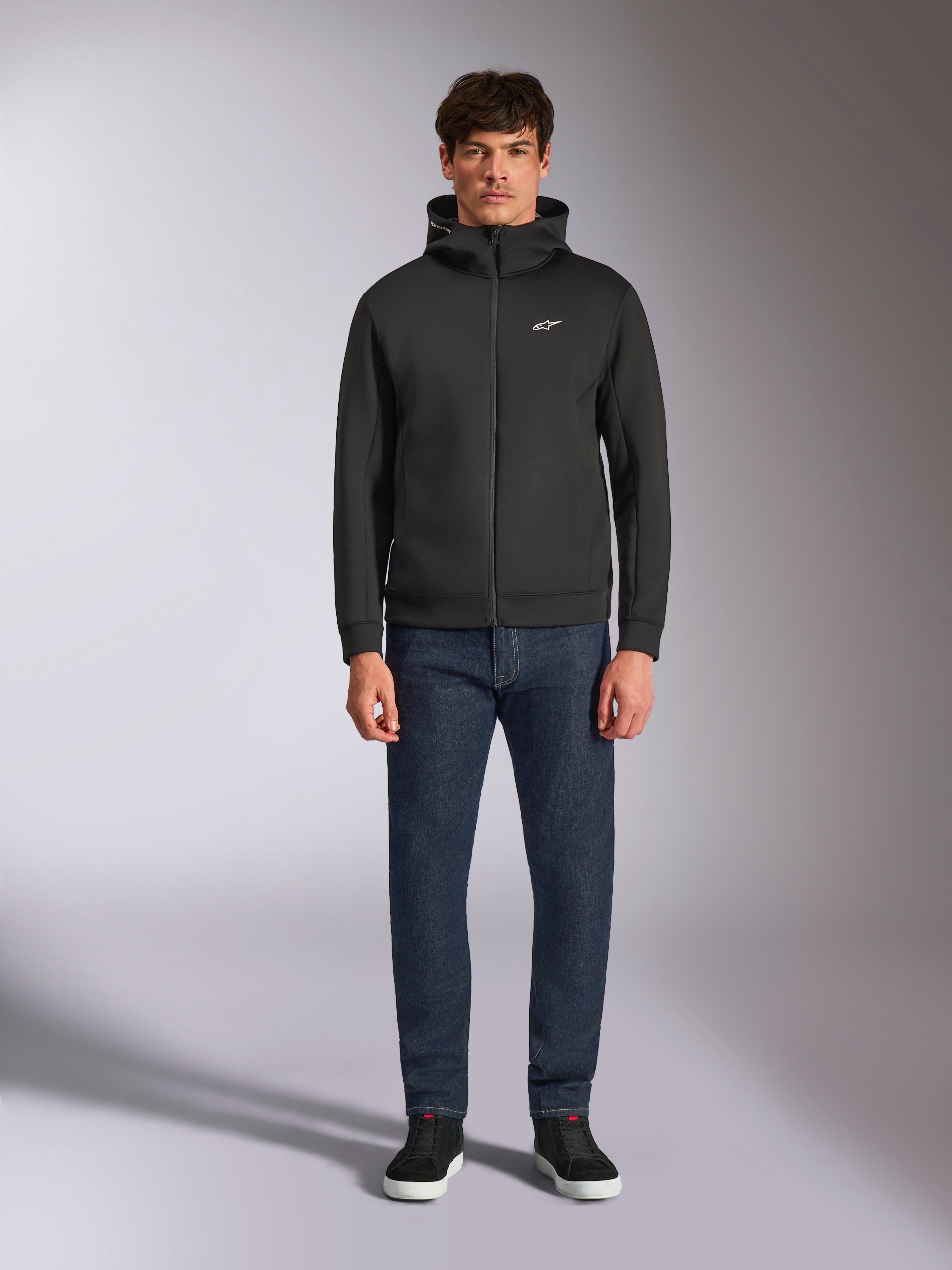 Model w bluzie Alpinestars Cypher Hoodie, czarna rozpinana bluza z polaru, wysoki kaptur z małym białym logo Astars na piersi, ciemne jeansy i czarne sneakersy, casualowy streetwear inspirowany motorsportem