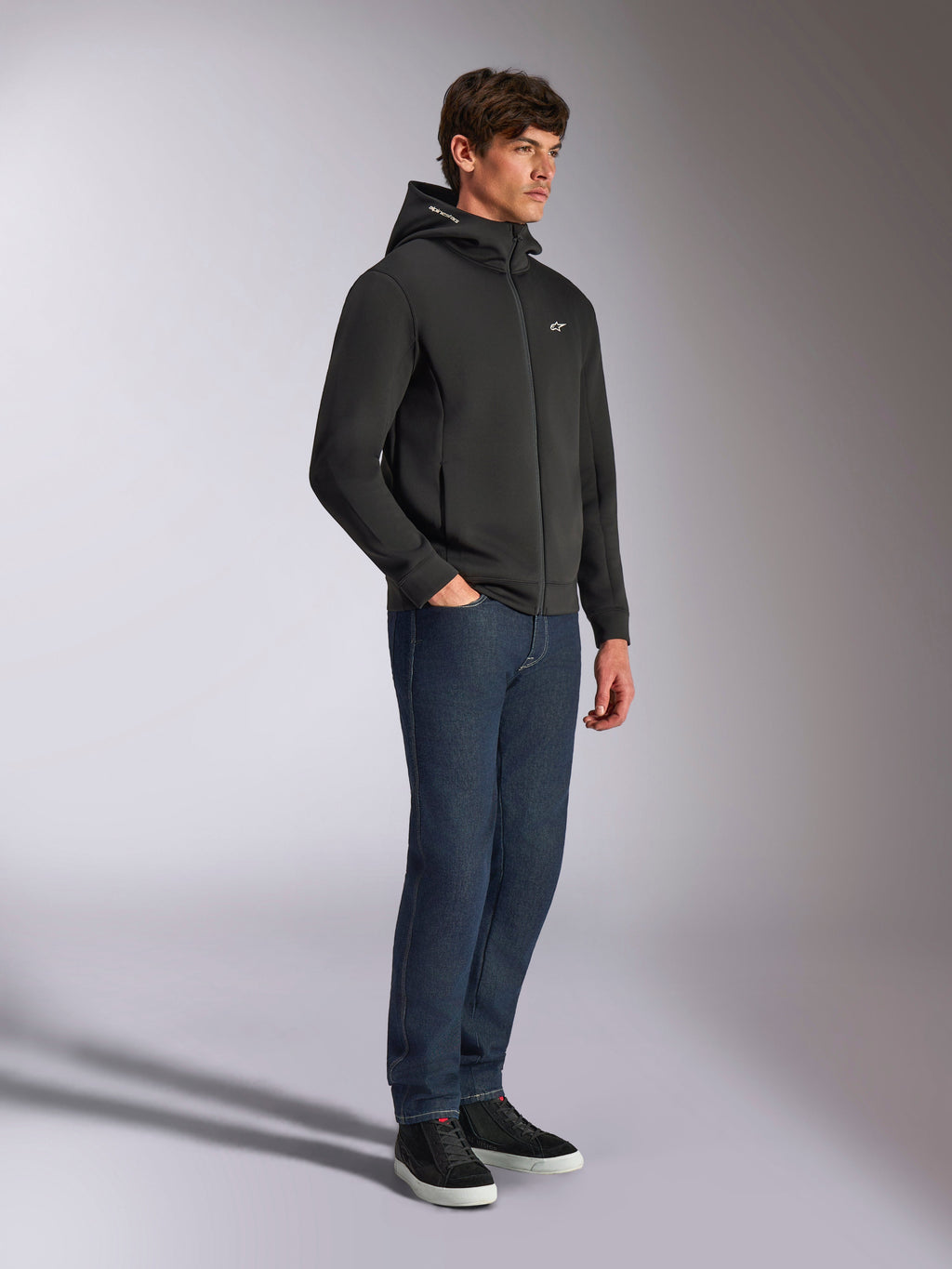 Alpinestars Cypher Hoodie, casual streetwear, czarna bluza z kapturem zapinana na zamek, męski model ubrany w ciemnoniebieskie dżinsy i czarne sneakersy, wysoki kaptur z logotypem Alpinestars, małe białe logo Astars na piersi, stylowy wygląd lifestylowy inspirowany sportami motorowymi