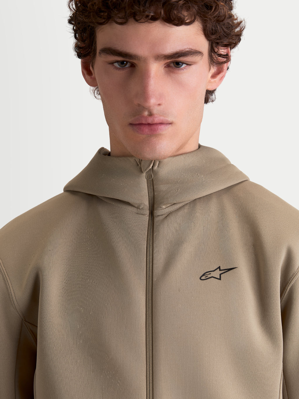 Alpinestars bluza z kapturem Cypher, beżowa sportowa bluza z wysokim kapturem prezentowana na modelu, pełny zamek, małe czarne logo Alpinestars na piersi, techniczna tkanina tri-blend, codzienna odzież sportowa
