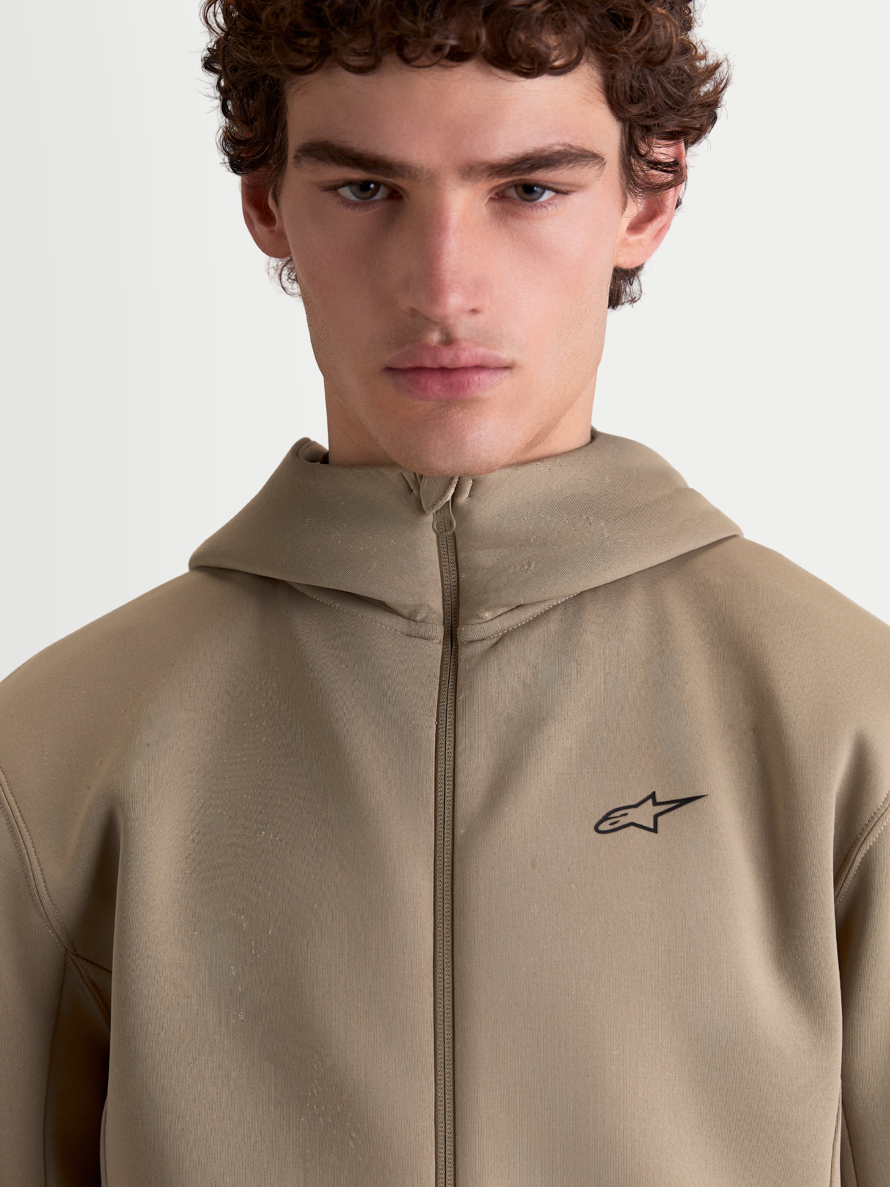 Alpinestars bluza z kapturem Cypher, beżowa sportowa bluza z wysokim kapturem prezentowana na modelu, pełny zamek, małe czarne logo Alpinestars na piersi, techniczna tkanina tri-blend, codzienna odzież sportowa