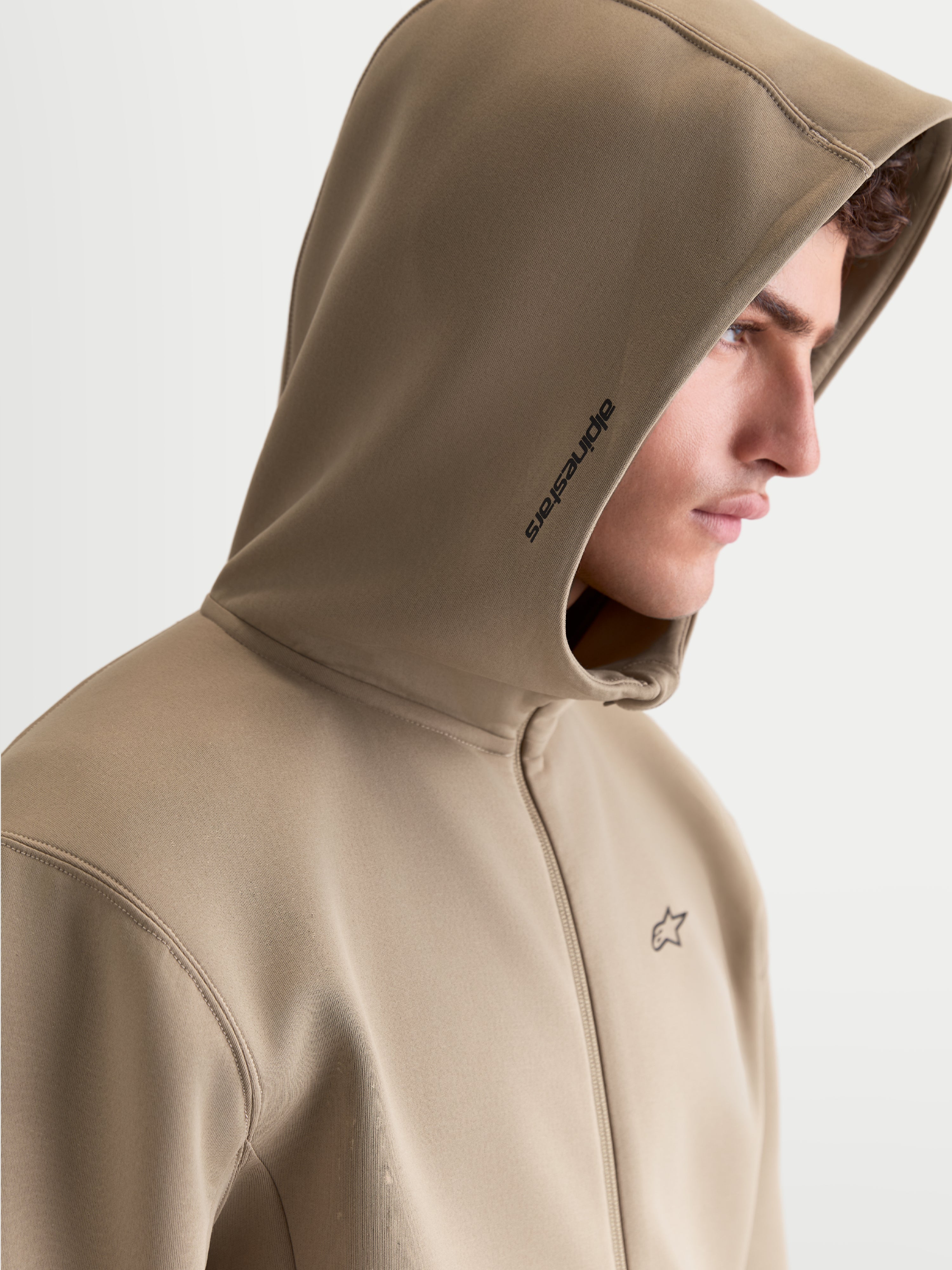 Alpinestars bluza z kapturem Cypher, męska beżowa bluza polarowa zapinana na zamek, wysoki kołnierz z kapturem z siatkową podszewką i brandingiem Alpinestars na boku, małe logo na klatce piersiowej, widok z profilu na modelu, odzież sportowa w stylu motorsport.