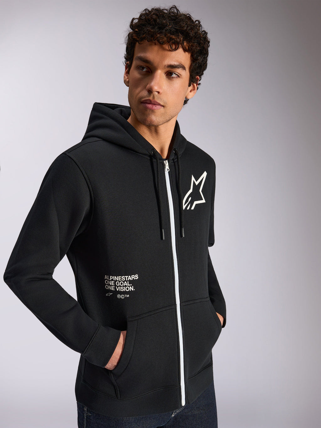 Bluza z kapturem Alpinestars Transverse, odzież sportowa typu casual, czarna z białym detalem zamka, kaptur ze sznurkami, logo gwiazdy Alpinestars na piersi, grafika „One Goal One Vision”, męski model z ciemnymi, kręconymi włosami