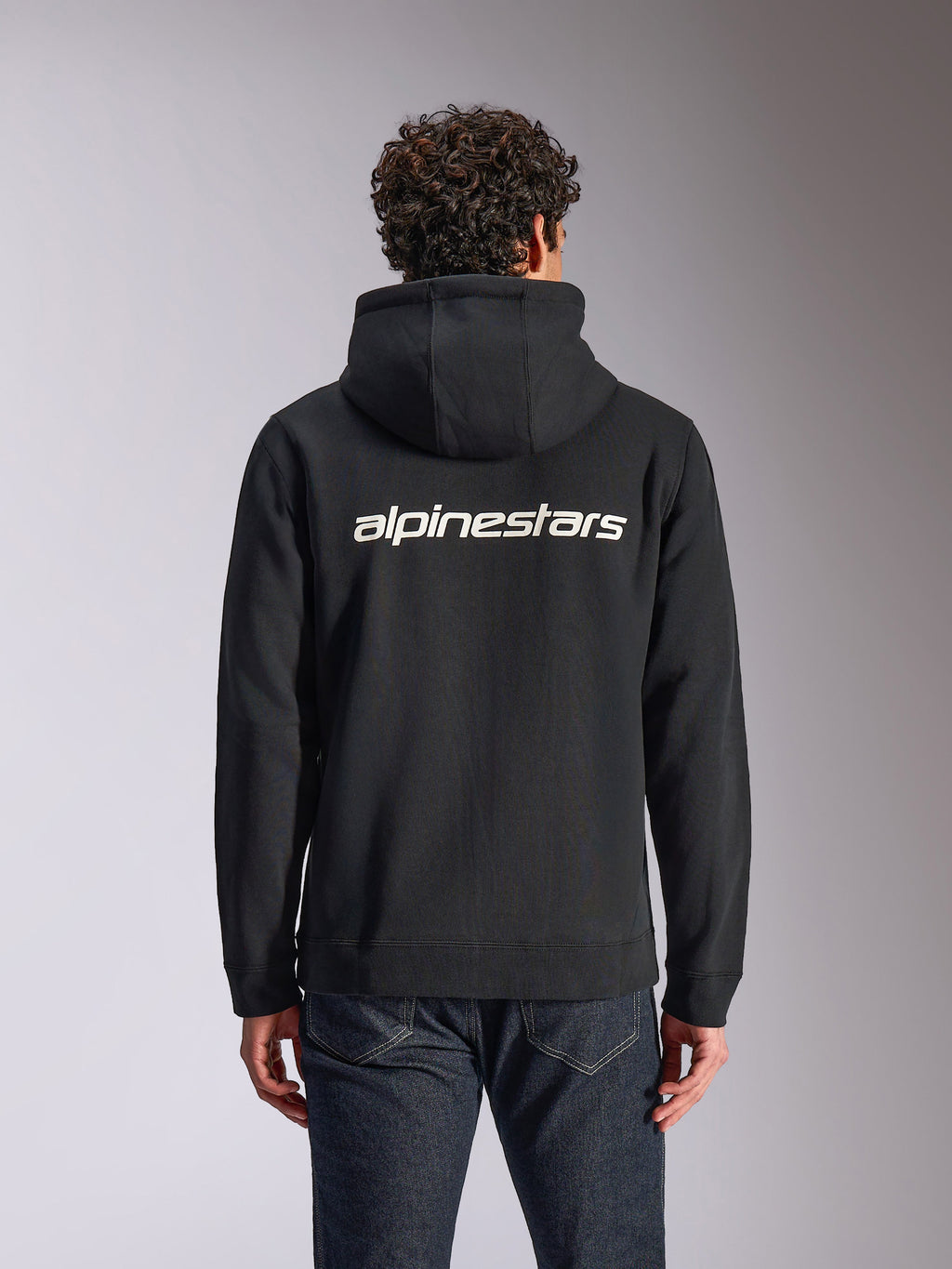 Alpinestars Transverse Hoodie, casualowa odzież streetwear, czarna z białym logotypem Alpinestars na plecach, rozpinana bluza z polaru z kapturem, długie rękawy, noszona przez modela do ciemnych jeansów, nowoczesny design inspirowany motorsportem