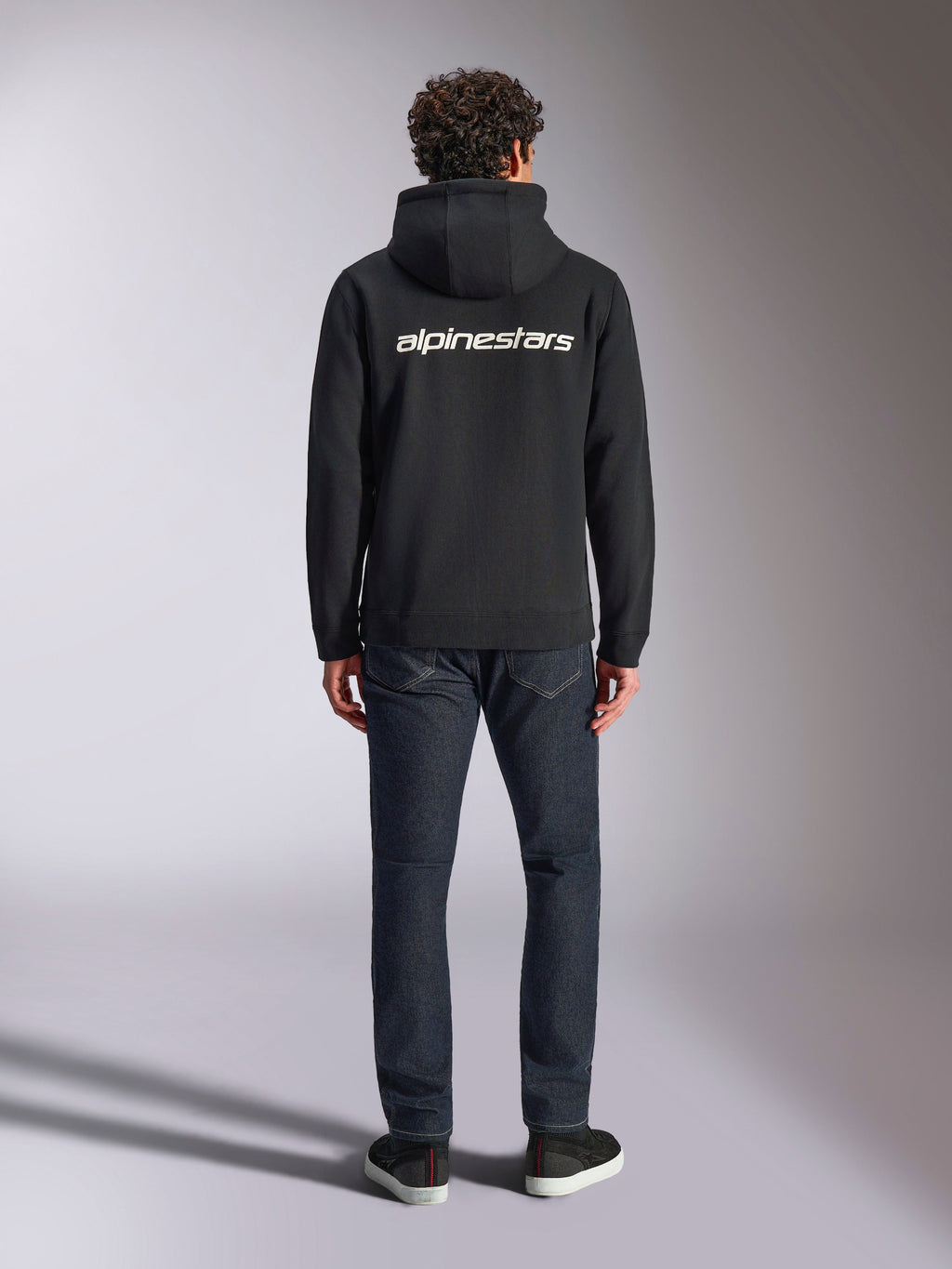 Bluza z kapturem Alpinestars Transverse, codzienna odzież streetwearowa w stylu motorsport, czarna z białym logotypem Alpinestars na plecach, długie rękawy, prążkowany dół i mankiety, widok z tyłu w zestawieniu z ciemnymi dżinsami