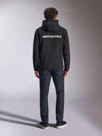Bluza z kapturem Alpinestars Transverse, codzienna odzież streetwearowa w stylu motorsport, czarna z białym logotypem Alpinestars na plecach, długie rękawy, prążkowany dół i mankiety, widok z tyłu w zestawieniu z ciemnymi dżinsami