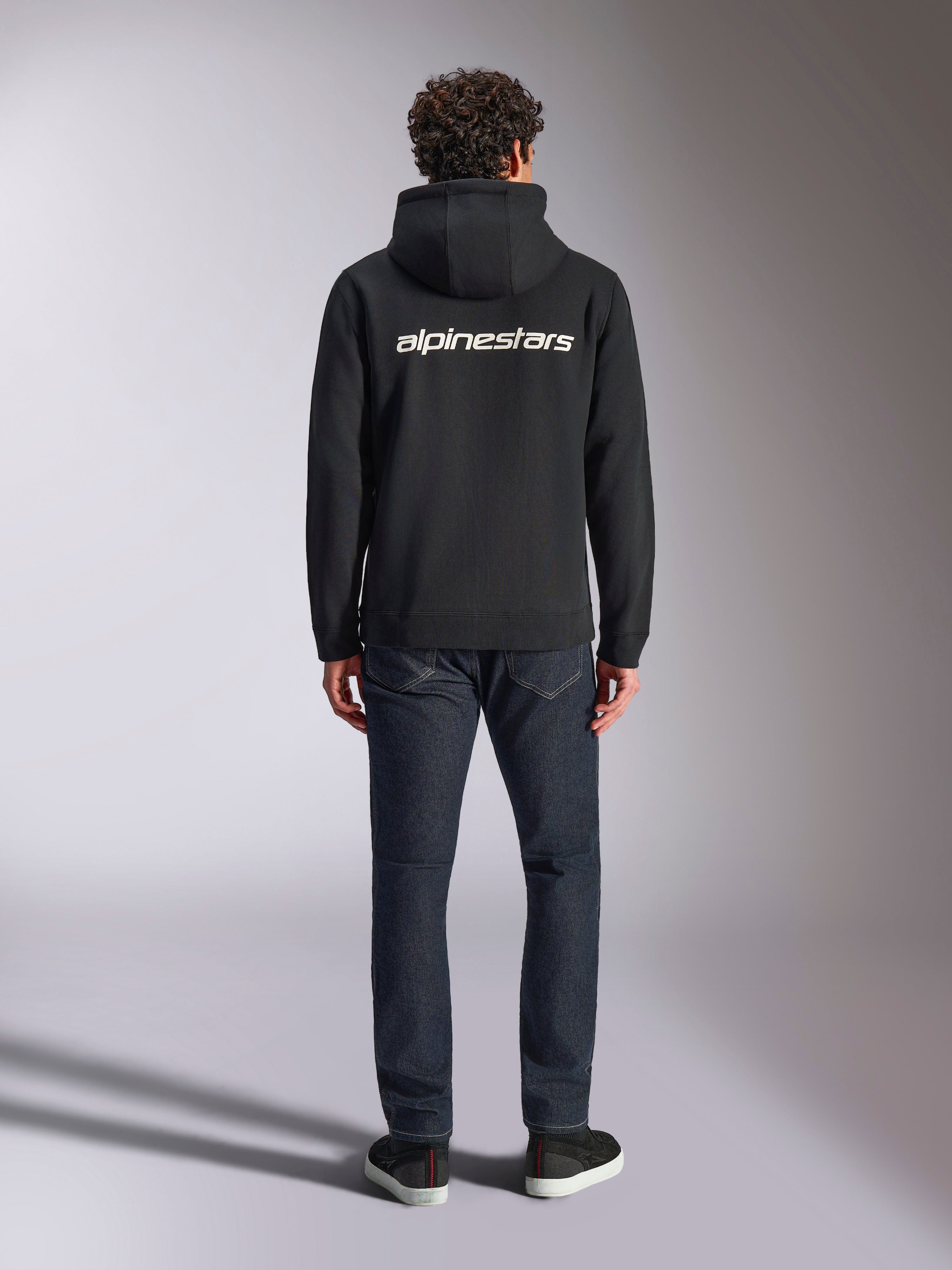 Bluza z kapturem Alpinestars Transverse, codzienna odzież streetwearowa w stylu motorsport, czarna z białym logotypem Alpinestars na plecach, długie rękawy, prążkowany dół i mankiety, widok z tyłu w zestawieniu z ciemnymi dżinsami