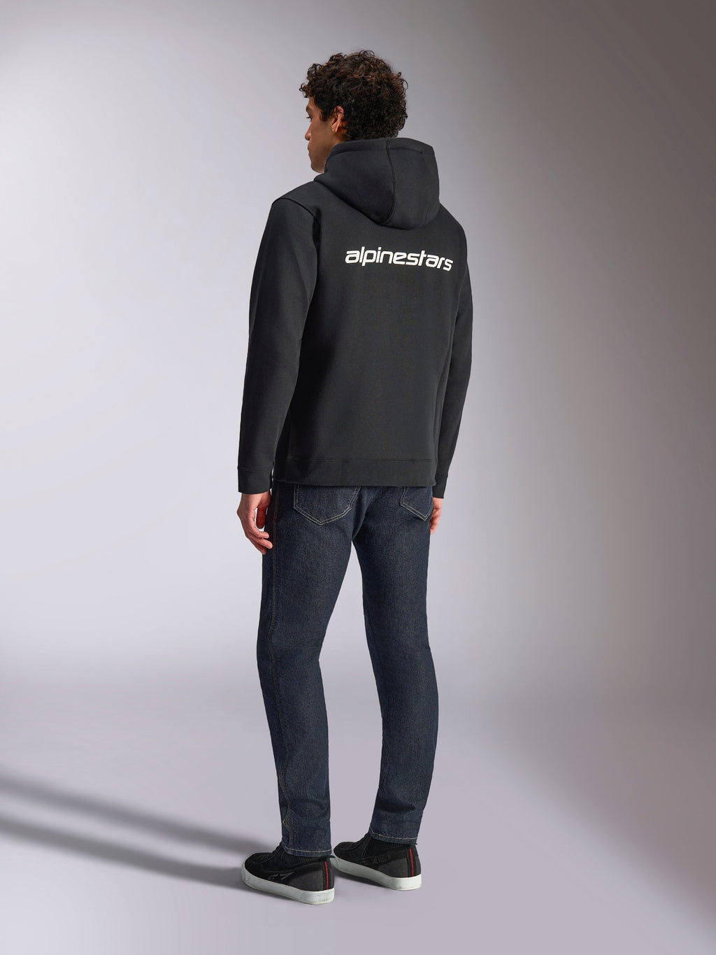 Alpinestars Transverse Hoodie, czarna rozpinana bluza z polaru, widok z tyłu z białym logotypem Alpinestars na plecach, zestawiona z ciemnoniebieskimi dżinsami i czarnymi sneakersami, codzienna odzież streetwearowa inspirowana sportami motorowymi