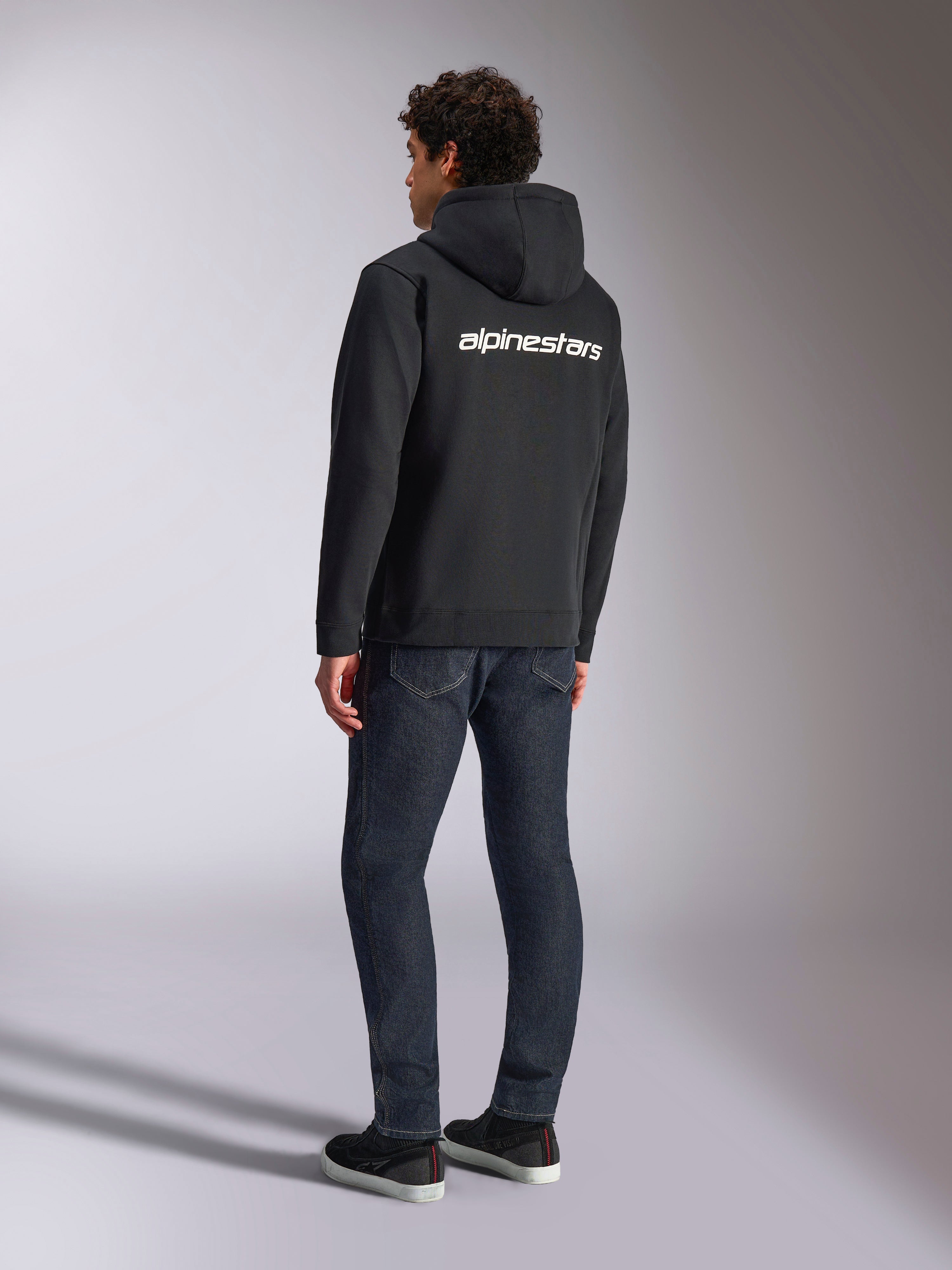 Alpinestars Transverse Hoodie, czarna rozpinana bluza z polaru, widok z tyłu z białym logotypem Alpinestars na plecach, zestawiona z ciemnoniebieskimi dżinsami i czarnymi sneakersami, codzienna odzież streetwearowa inspirowana sportami motorowymi