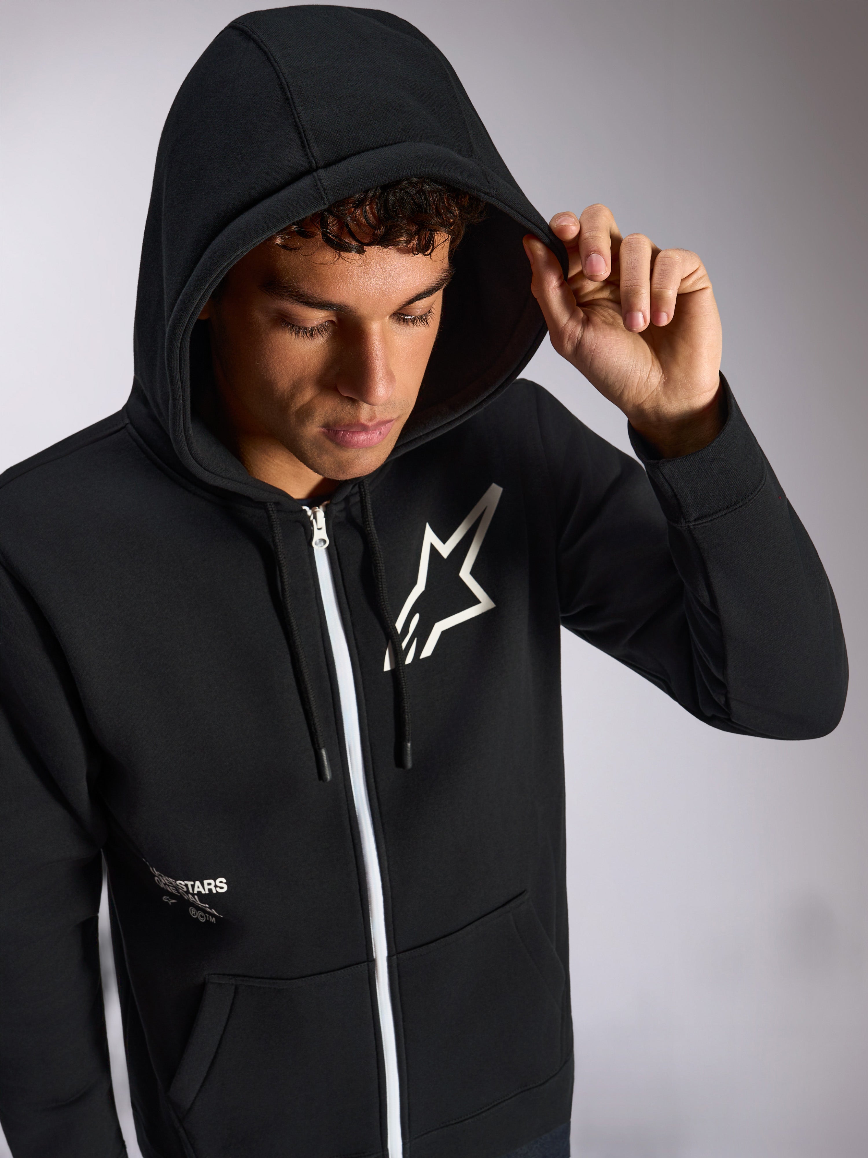Alpinestars Transverse Hoodie, czarna bluza rozpinana z kapturem, białe logo Astars na piersi, biały zamek błyskawiczny, założony kaptur ze ściągaczem, codzienna odzież uliczna inspirowana sportami motorowymi