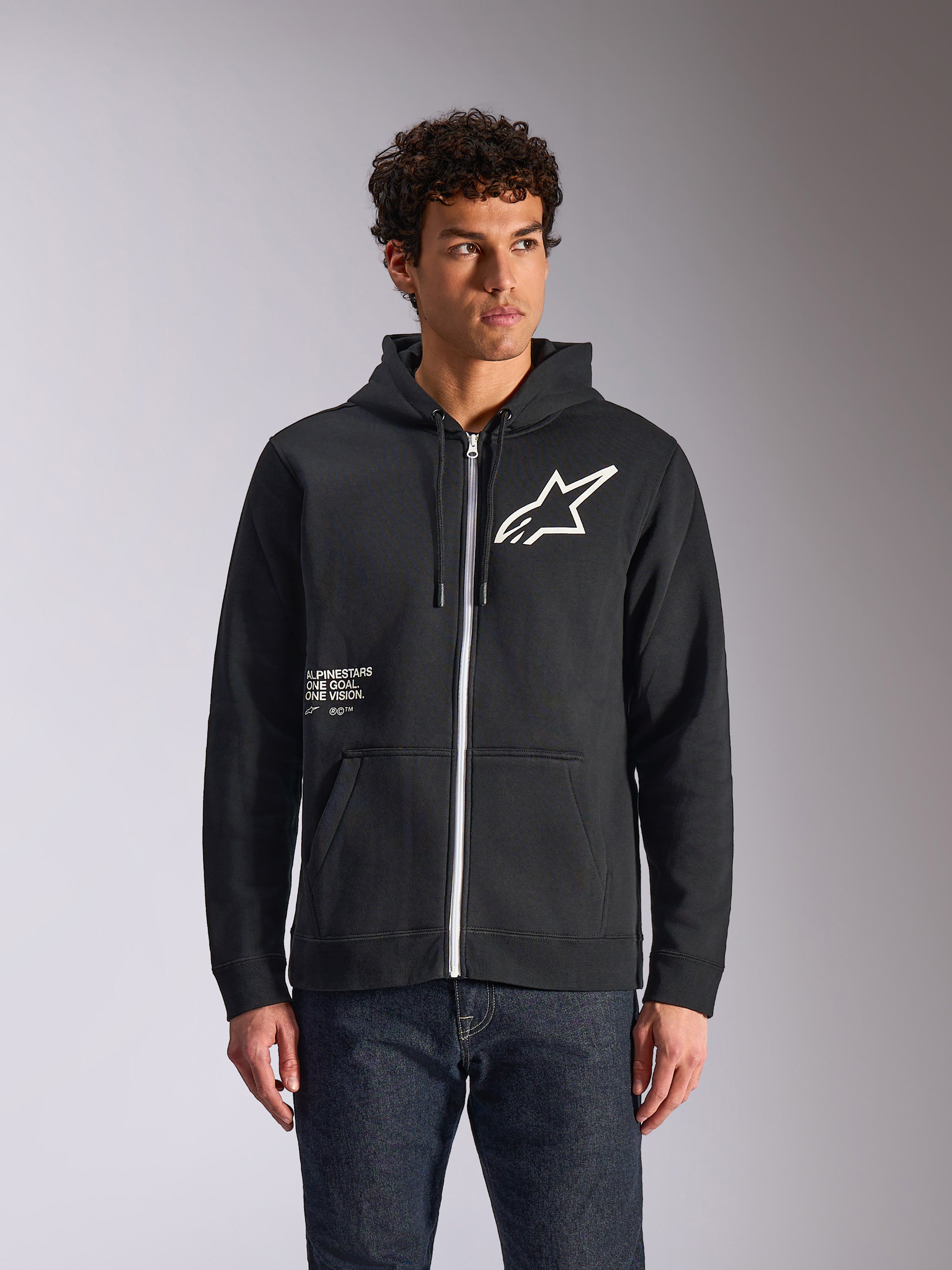 Alpinestars Transverse Hoodie, czarna bluza z kapturem zapinana na zamek, białe logo Astars i grafika One Goal z przodu, model w ciemnych dżinsach, codzienna odzież uliczna inspirowana sportami motorowymi