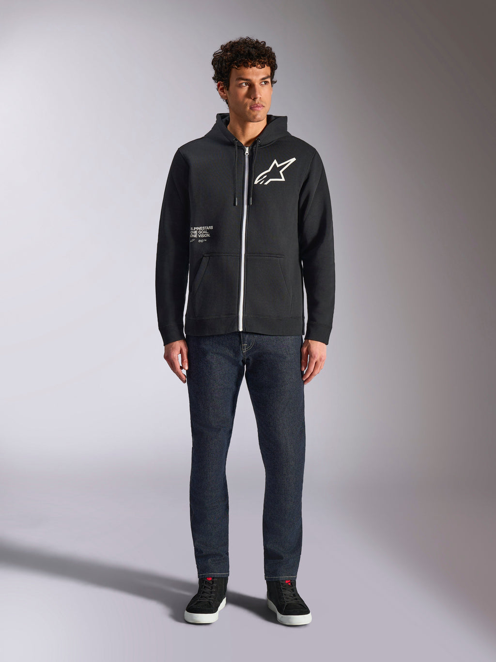 Alpinestars Transverse Hoodie, codzienna bluza polarowa rozpinana z przodu, czarna, białe logo Astars na piersi i nadruk „One Goal” z przodu, kaptur ze sznurkiem, w zestawieniu z ciemnymi jeansami i czarnymi sneakersami, lifestyle'owa odzież uliczna w stylu motorsport