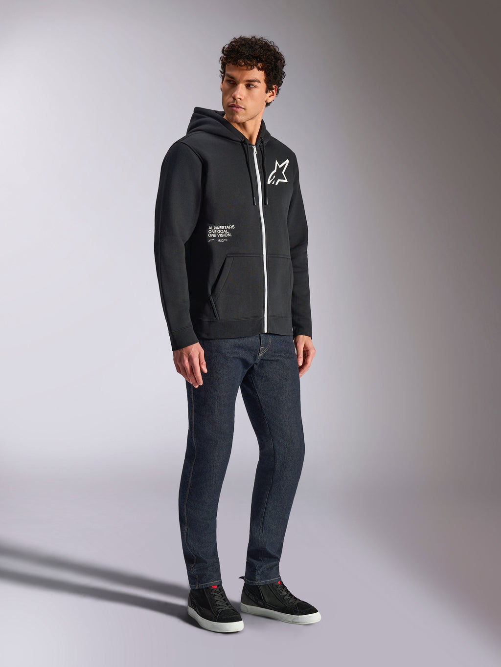 Alpinestars Transverse Hoodie, czarna rozpinana bluza z kapturem z kontrastowym białym zamkiem, białe logo gwiazdy na piersi i napis „One Goal” na kieszeni, model w ciemnych jeansach i czarnych sneakersach, codzienna odzież typu streetwear inspirowana motorsportem