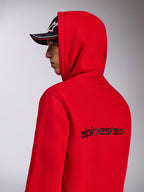 Alpinestars Transverse Hoodie, czerwona bluza z kapturem z polaru, widok z boku i z tyłu ukazujący czarny napis Alpinestars na plecach, model z założonym kapturem i czarną czapką z daszkiem, codzienna odzież uliczna inspirowana sportami motorowymi