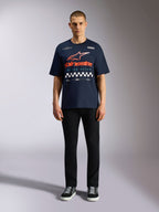 Alpinestars Aplomb Oversized CSF Tee, granatowy t-shirt z krótkim rękawem, duża grafika na klatce piersiowej z pomarańczowo-czerwonym gradientowym logo i wzorem szachownicy, model w czarnych spodniach i sneakersach, Casual Motorsport Streetwear