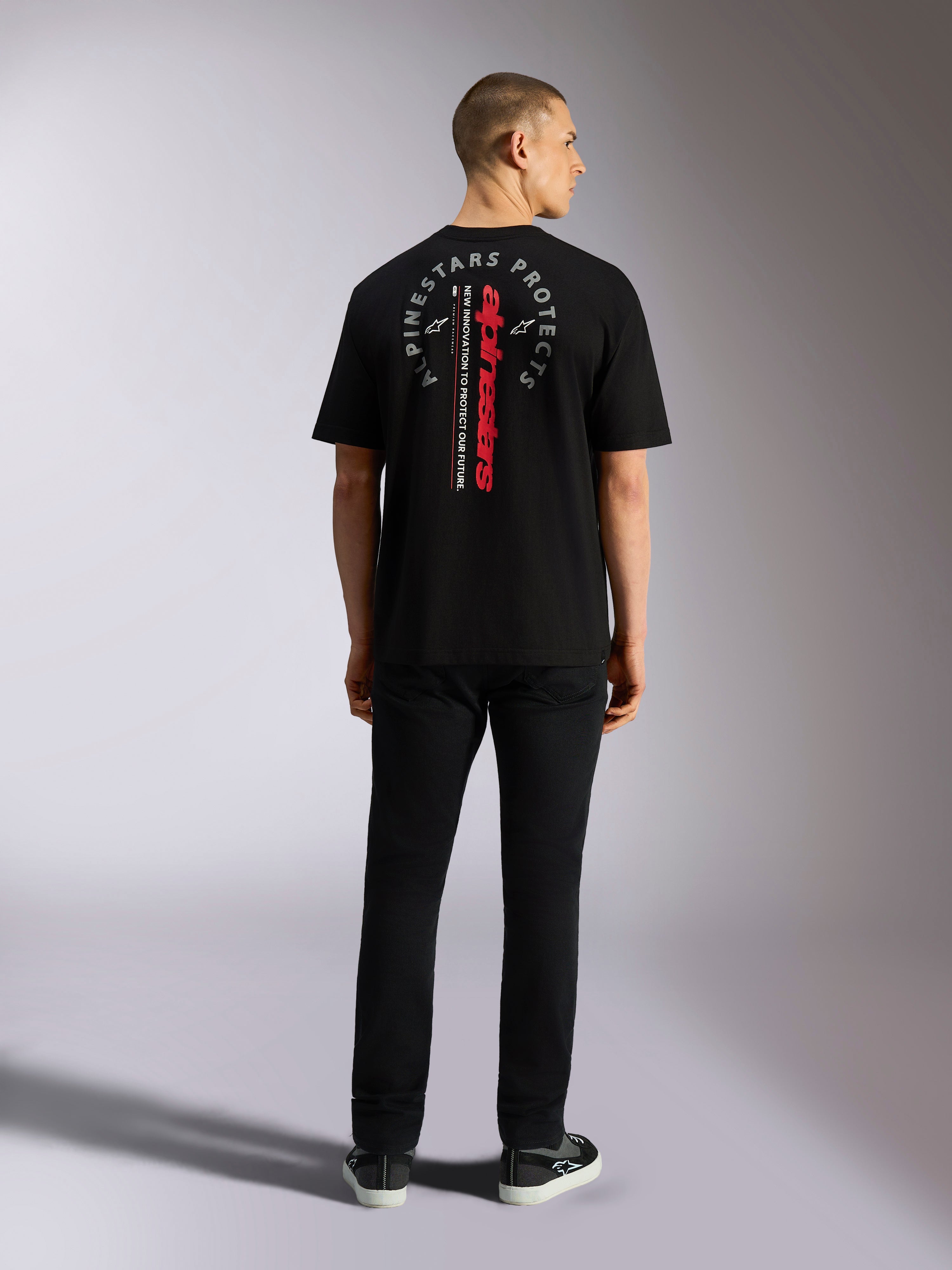 Alpinestars Overhang Oversized CSF Tee, koszulka z krótkim rękawem, czarna, z pionowym czerwonym napisem Alpinestars i szarą okrągłą grafiką na plecach, prezentowana przez modela od tyłu, codzienna odzież streetwearowa w stylu motorsportowym