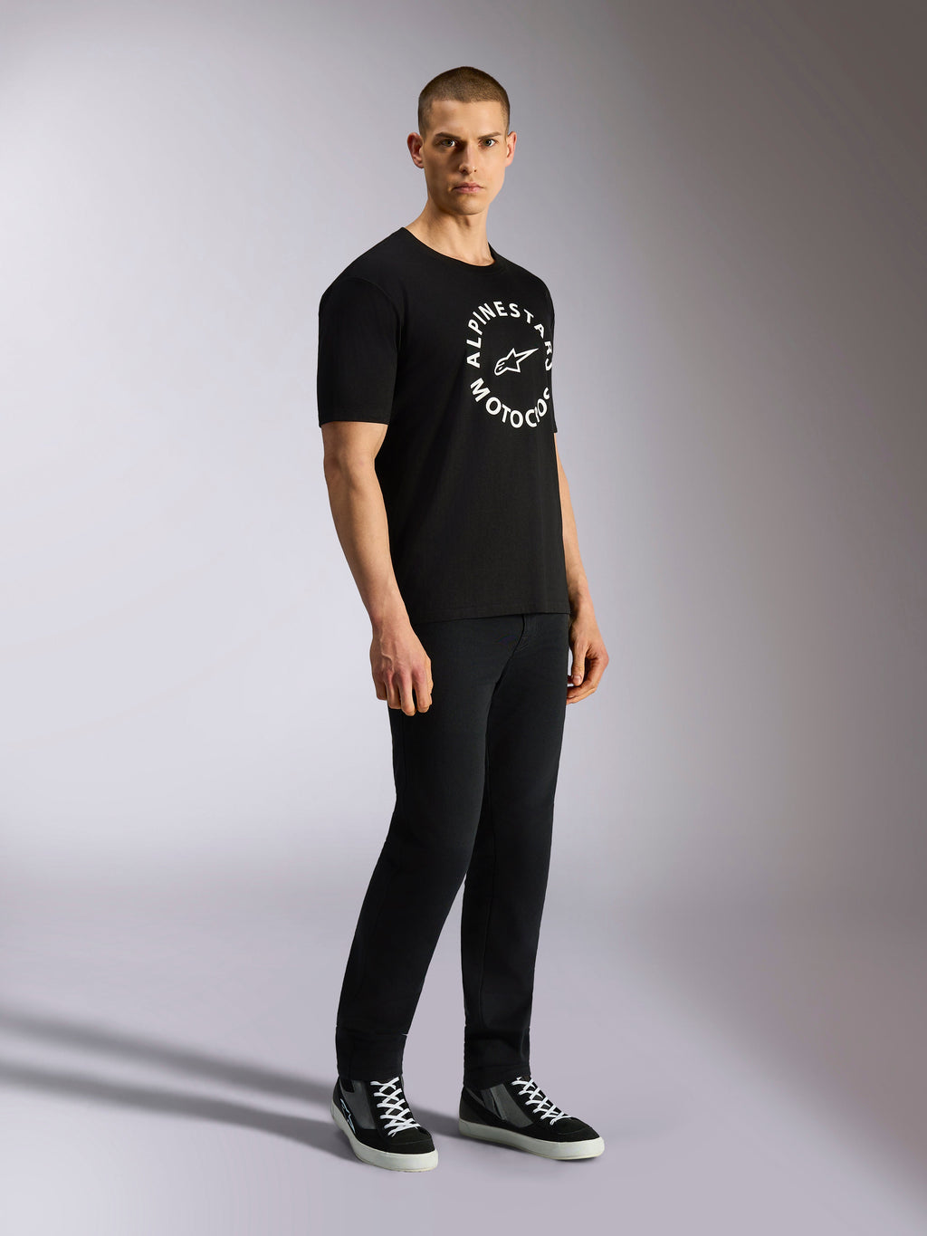 Koszulka Alpinestars MX CSF, T-shirt z krótkim rękawem, czarna, biała okrągła grafika Alpinestars Motocross na klatce piersiowej, noszona przez modela z czarnymi spodniami i tenisówkami, codzienny streetwear w stylu motorsportowym