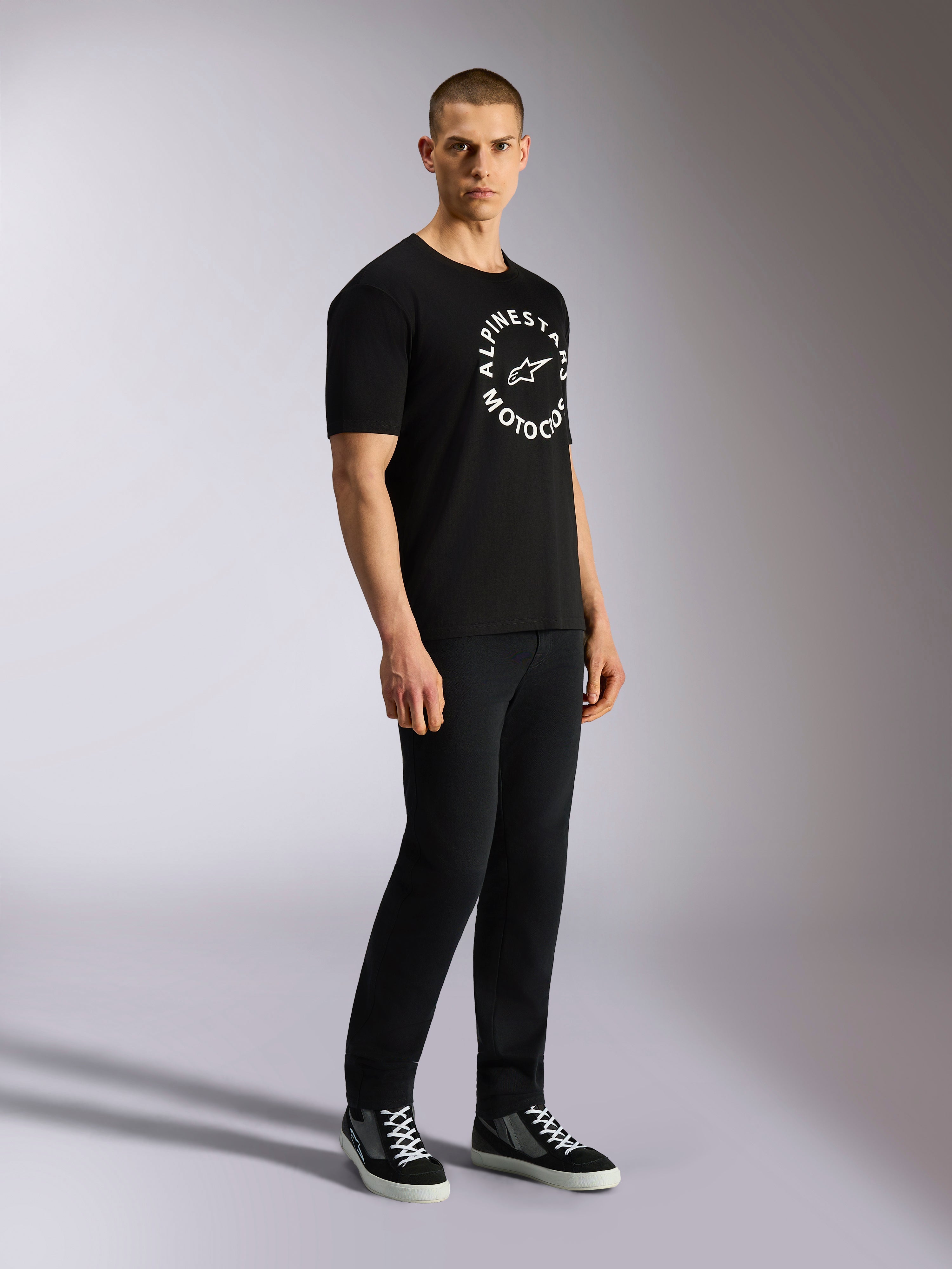 Koszulka Alpinestars MX CSF, T-shirt z krótkim rękawem, czarna, biała okrągła grafika Alpinestars Motocross na klatce piersiowej, noszona przez modela z czarnymi spodniami i tenisówkami, codzienny streetwear w stylu motorsportowym
