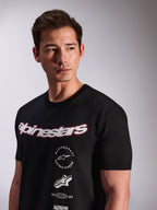 Alpinestars Abus CSF Tee, czarna koszulka z krótkim rękawem, noszona przez modela, białe logo Alpinestars z czerwoną obwódką i wielowarstwowe logotypy na klatce piersiowej, codzienna odzież streetwearowa w stylu motorsportowym